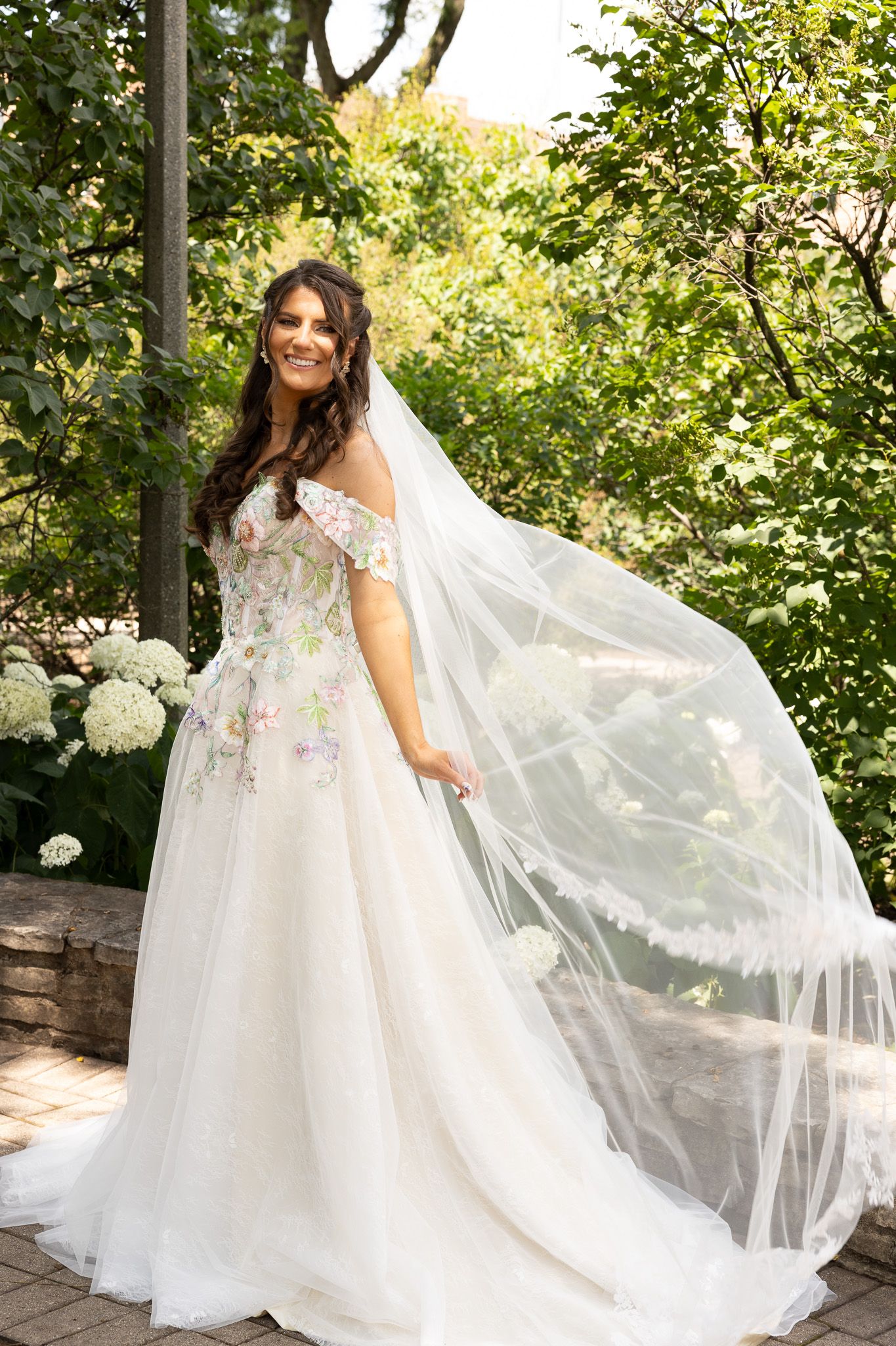 A bride in a flowing white gown with floral embellishments stands in a garden setting with a long veil trailing behind.