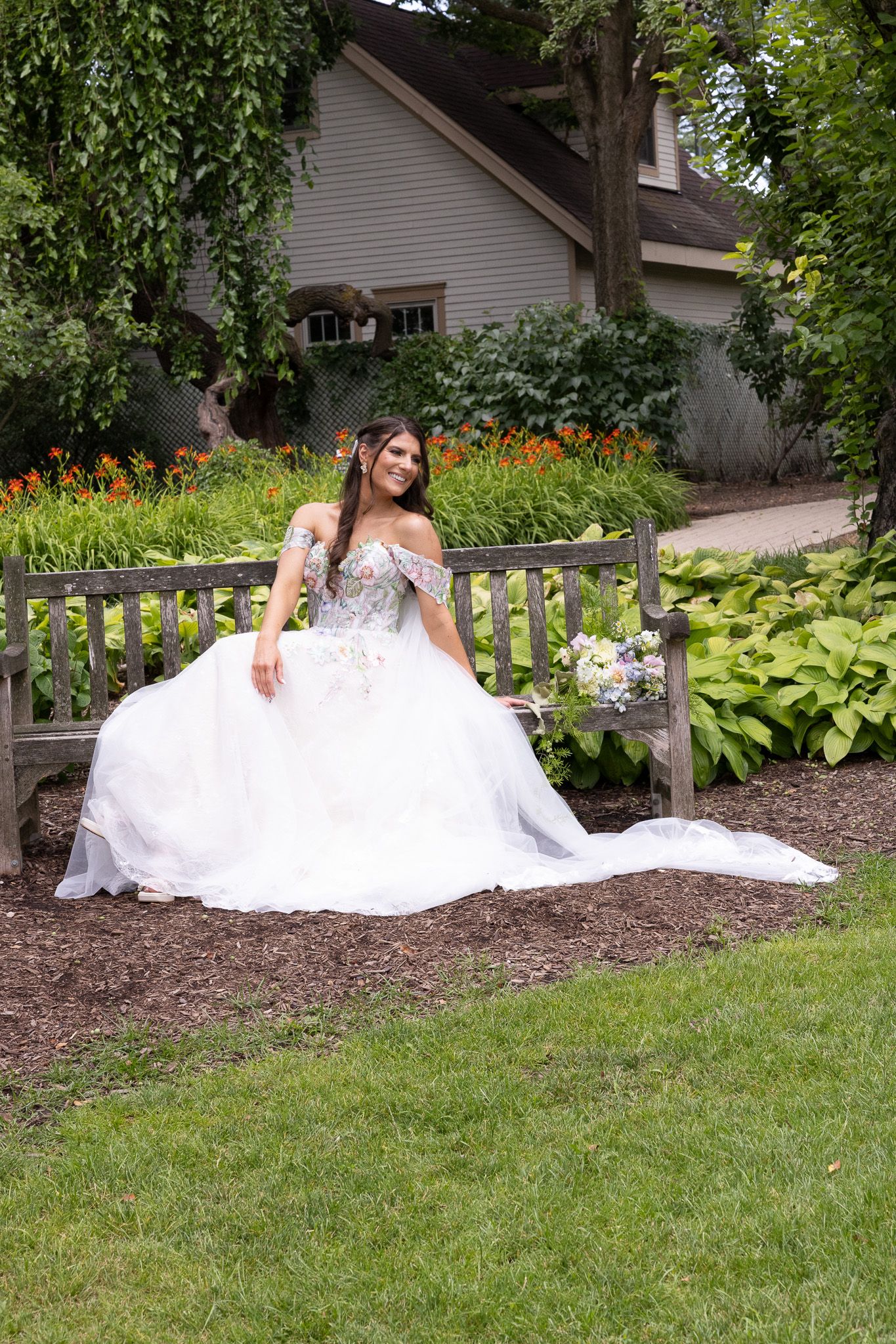 A bride in a white wedding dress poses elegantly on a garden bench surrounded by lush greenery and vibrant flowers.