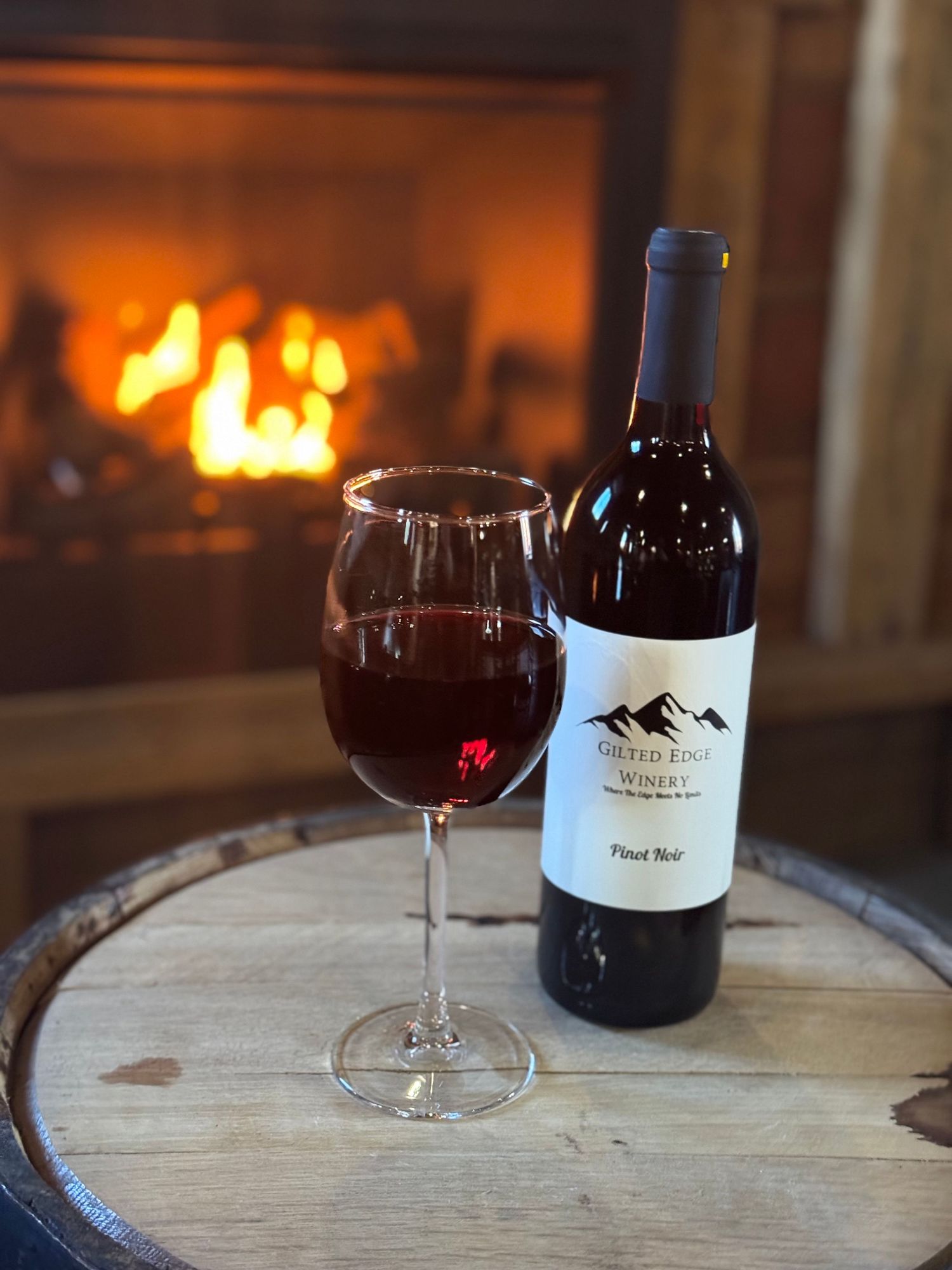 A glass and bottle of red wine sit on a wooden barrel in front of a glowing fireplace creating a cozy ambiance.
