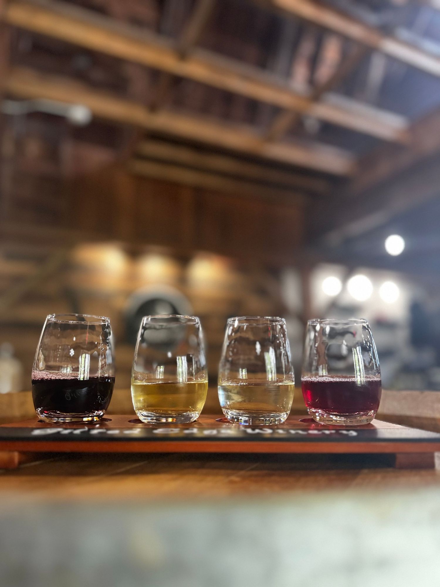 Four wine glasses containing different colored wines arranged on a wooden tasting flight board.