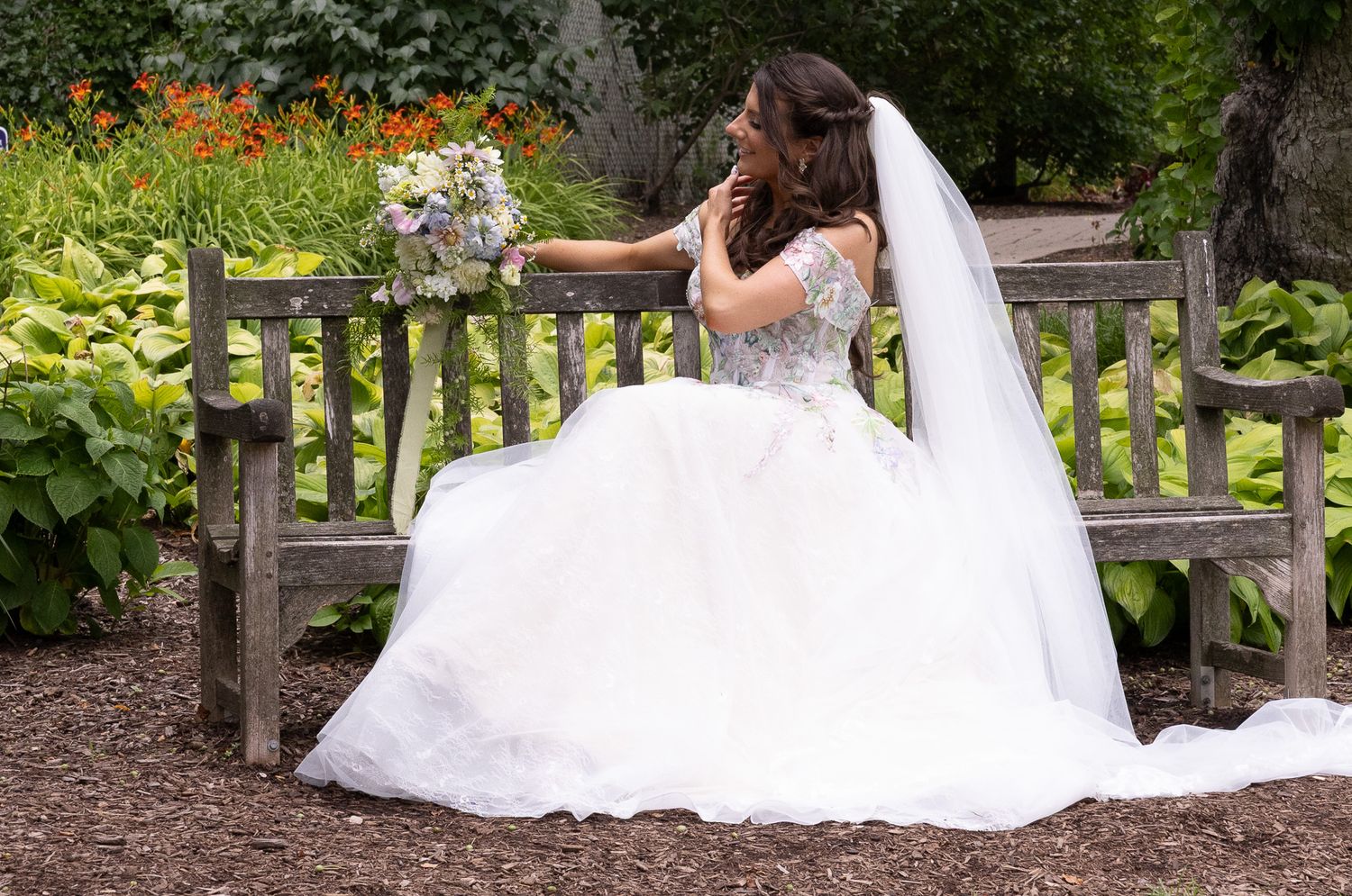A bride in an elegant white wedding dress sitting on a wooden garden bench in a lush outdoor setting.