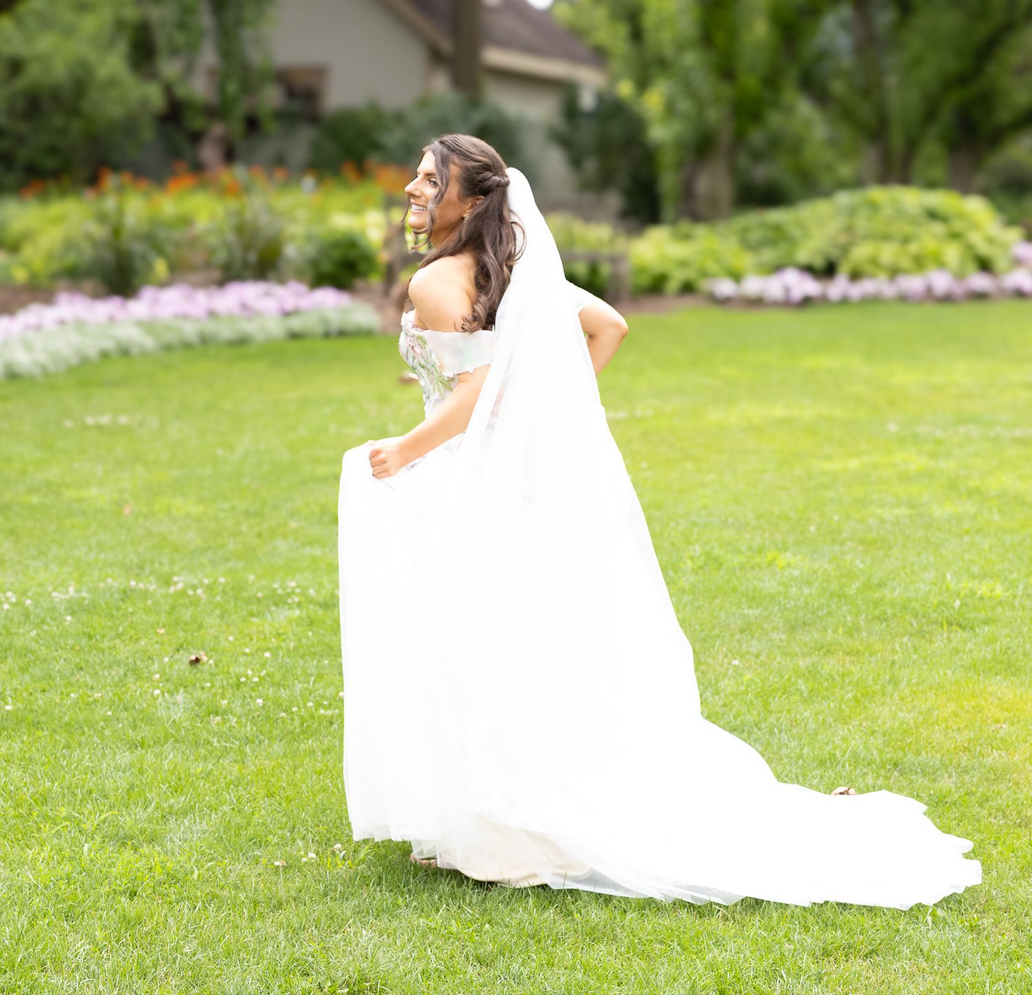 A bride in a flowing white gown reclines gracefully on lush green grass during a summer photoshoot.