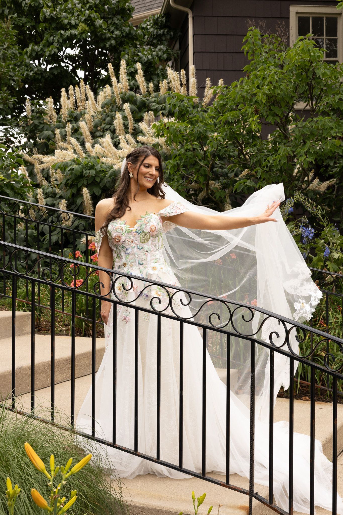 A white wedding dress flowing in a garden setting with ornate iron railing and blooming flowers.