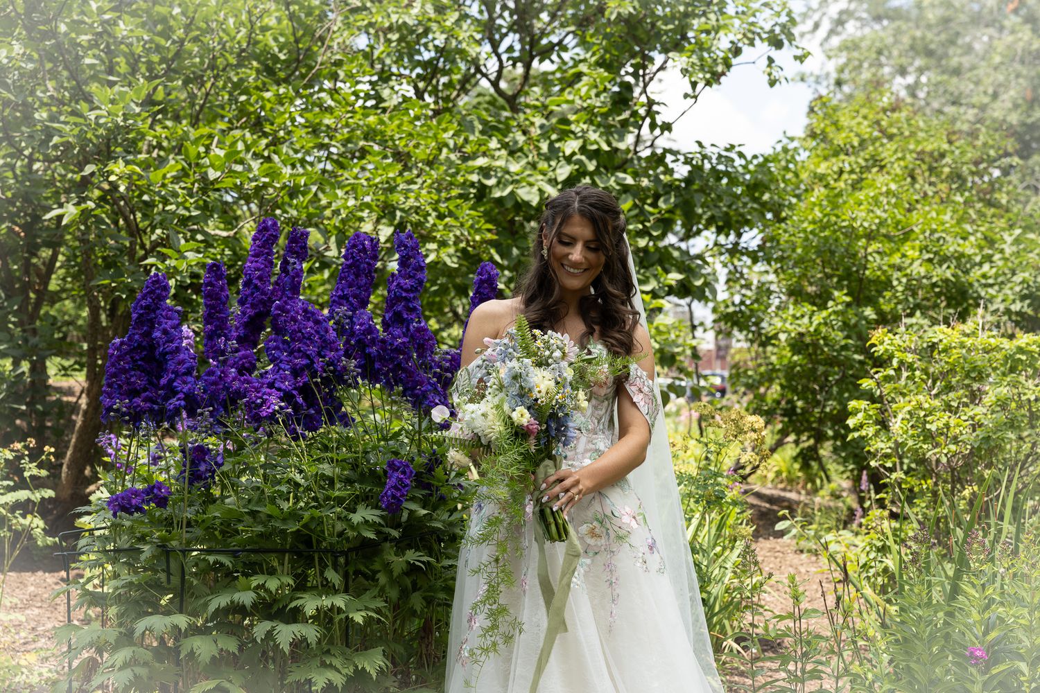 Beautiful outdoor garden wedding scene with bride holding bouquet and blue delphiniums blooming in the background.