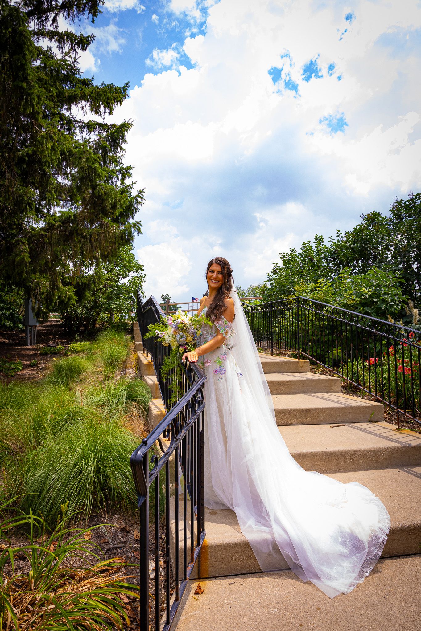 A flowing white wedding dress cascades down outdoor garden steps with lush greenery and blue sky in the background.
