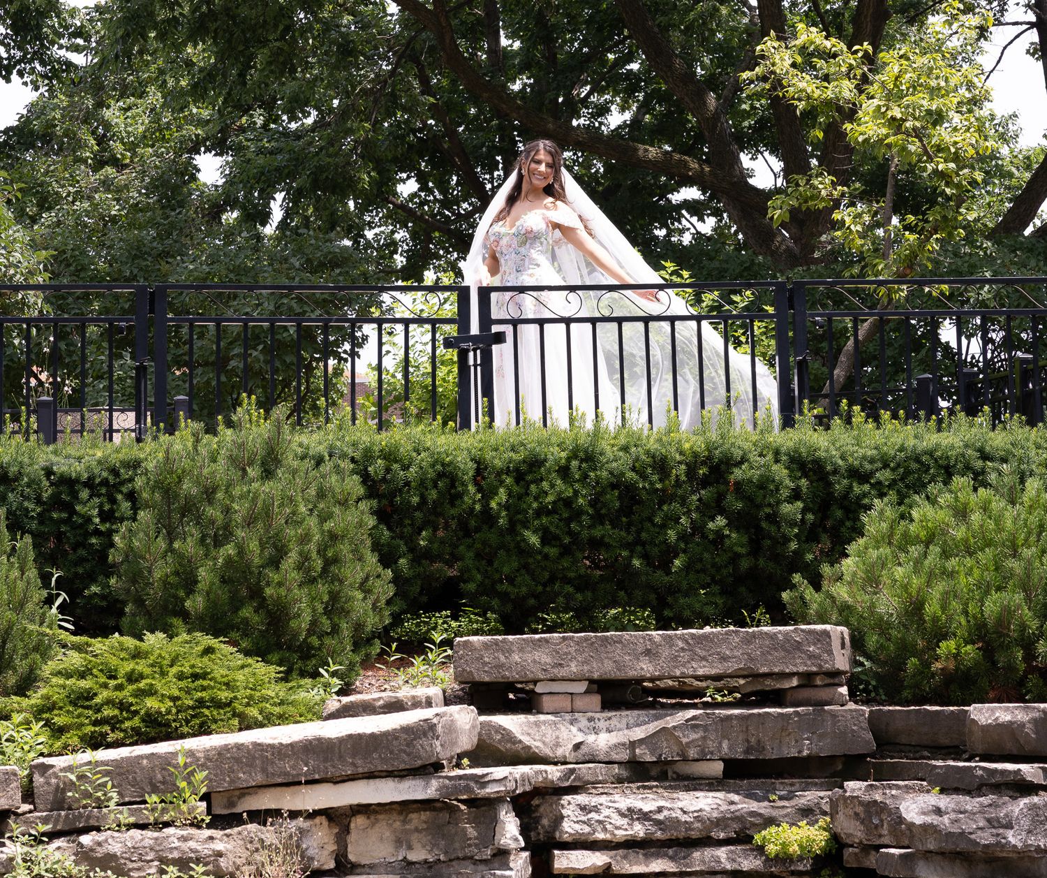 A panoramic view of a stone garden wall against lush greenery with wrought iron fencing in the background.