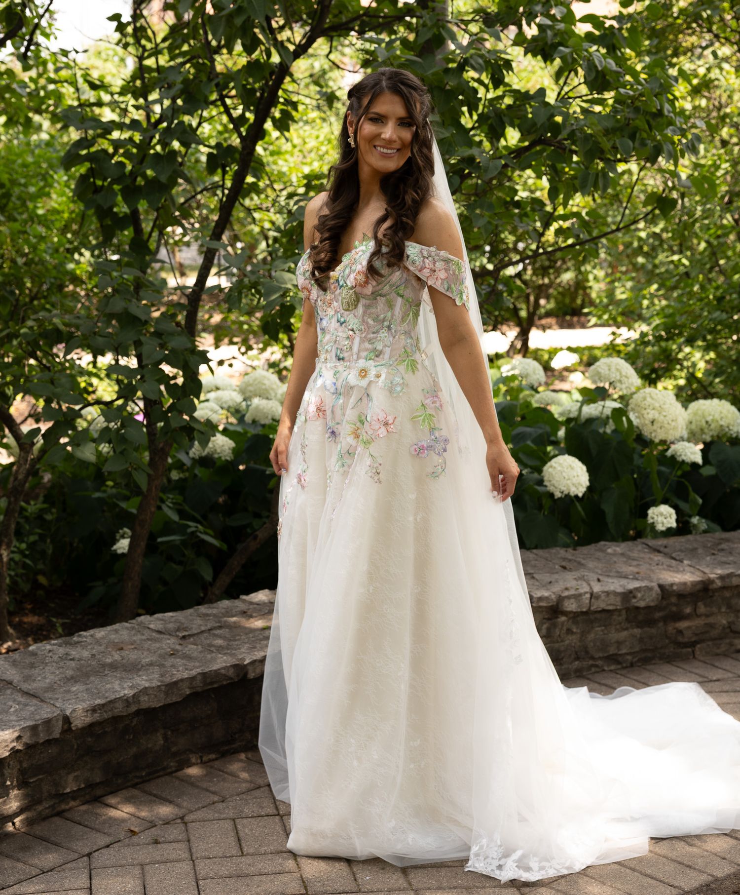 A bride in a flowing white tulle gown with floral embellishments poses on a stone path in a garden setting.