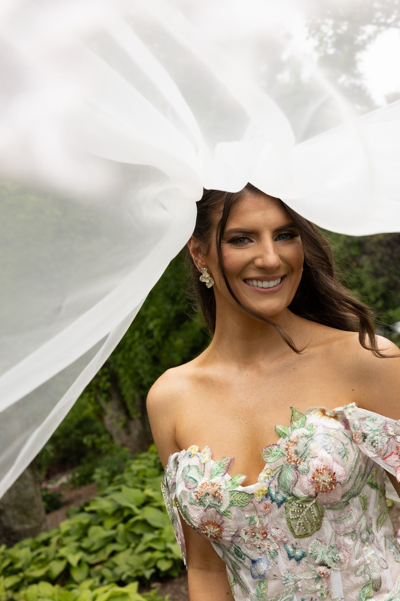 Wind sweeps through white veil above bride wearing strapless floral dress with sweetheart neckline in garden setting.