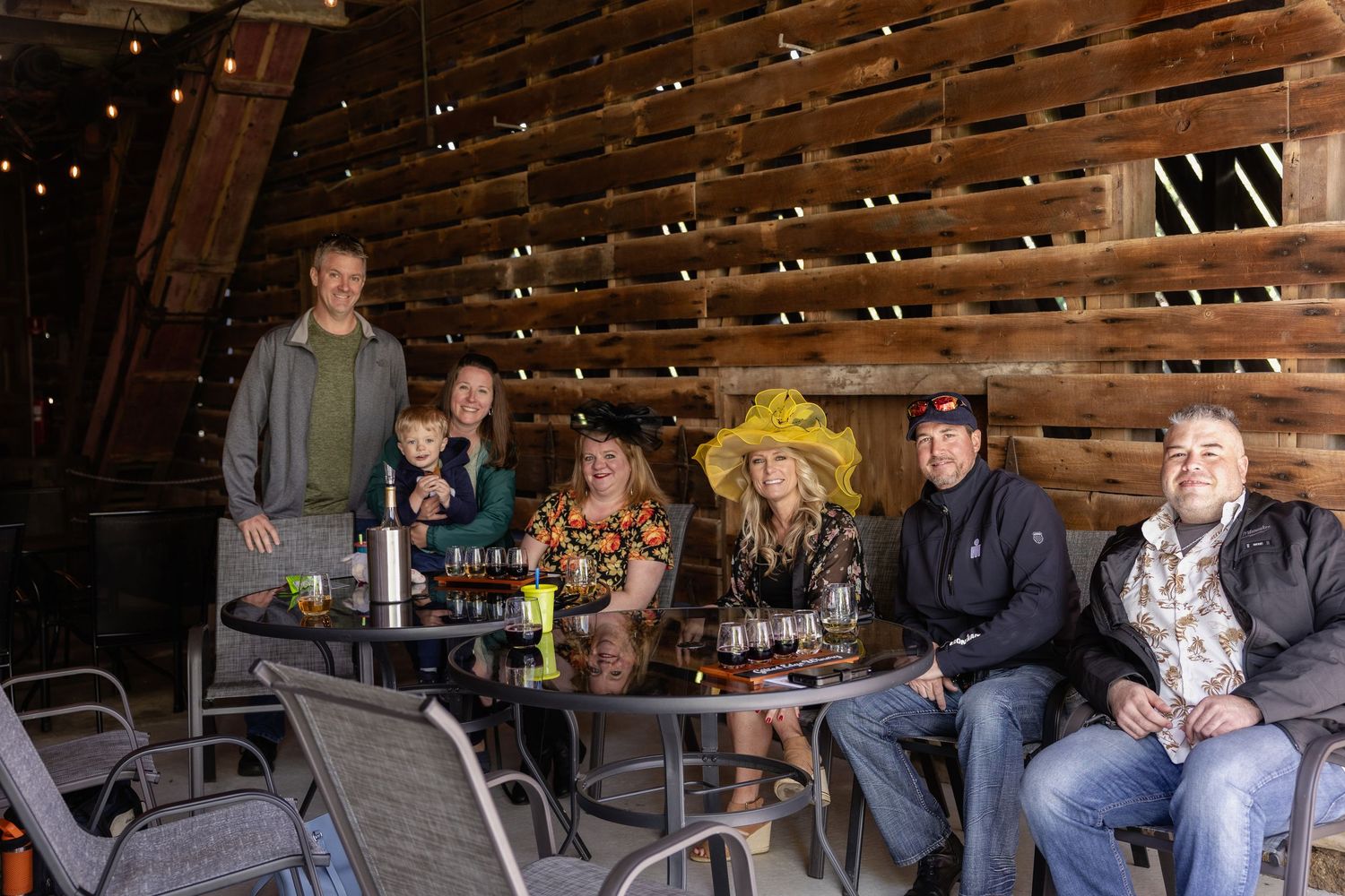 A group of people gather around a table on a covered rustic wooden patio with string lights overhead.