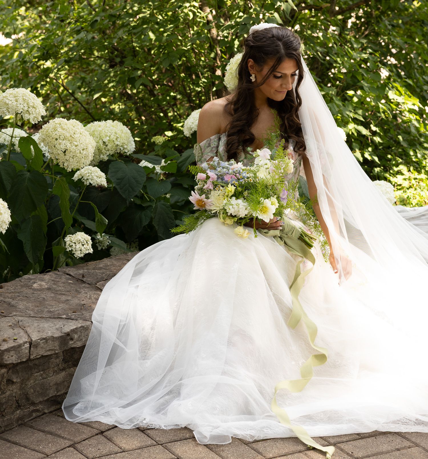 A bride in an elegant white gown sits gracefully on stone steps surrounded by lush hydrangeas and greenery in a garden.