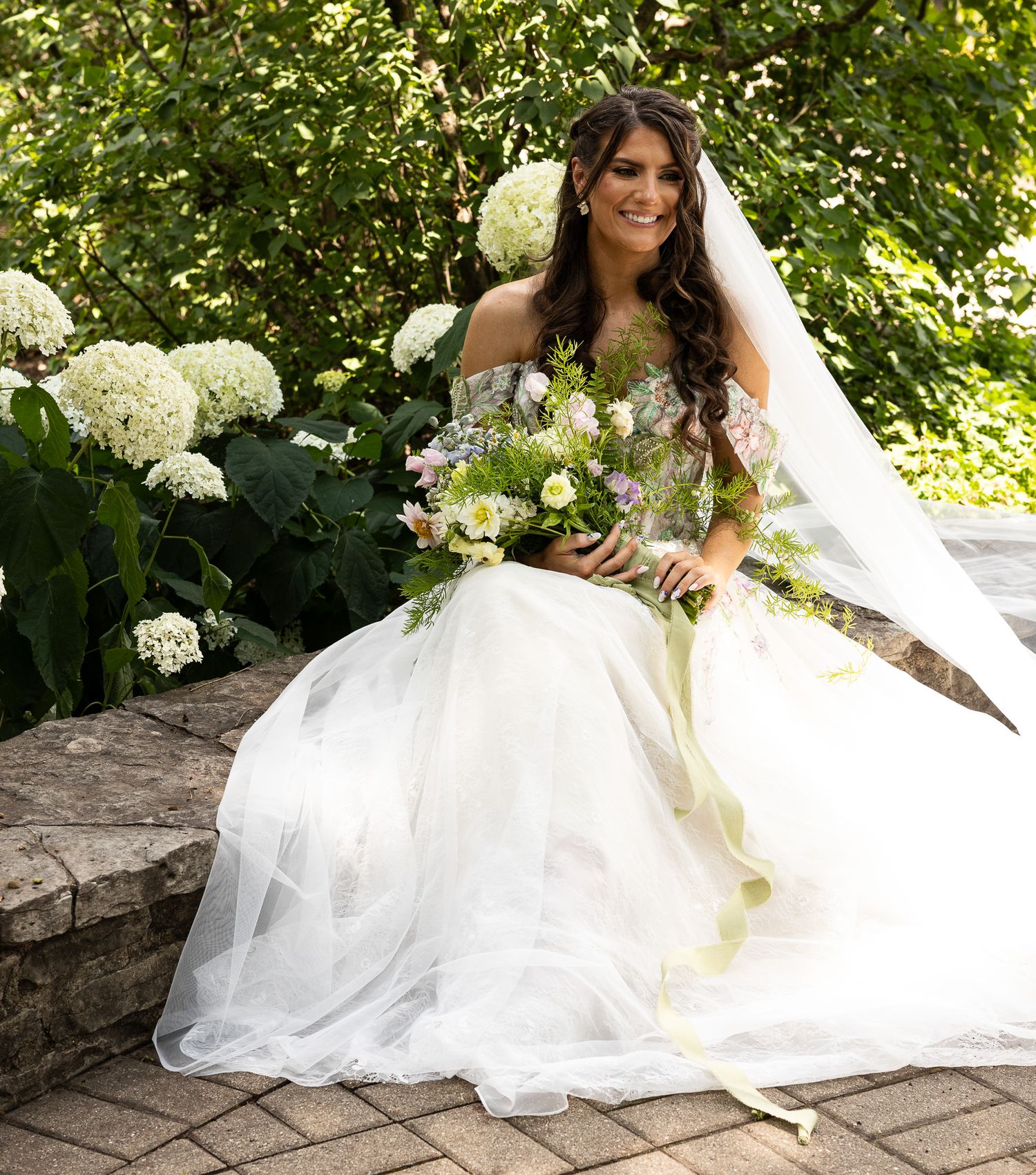 A bride in a white wedding gown poses gracefully on stone steps surrounded by blooming white hydrangeas and greenery.