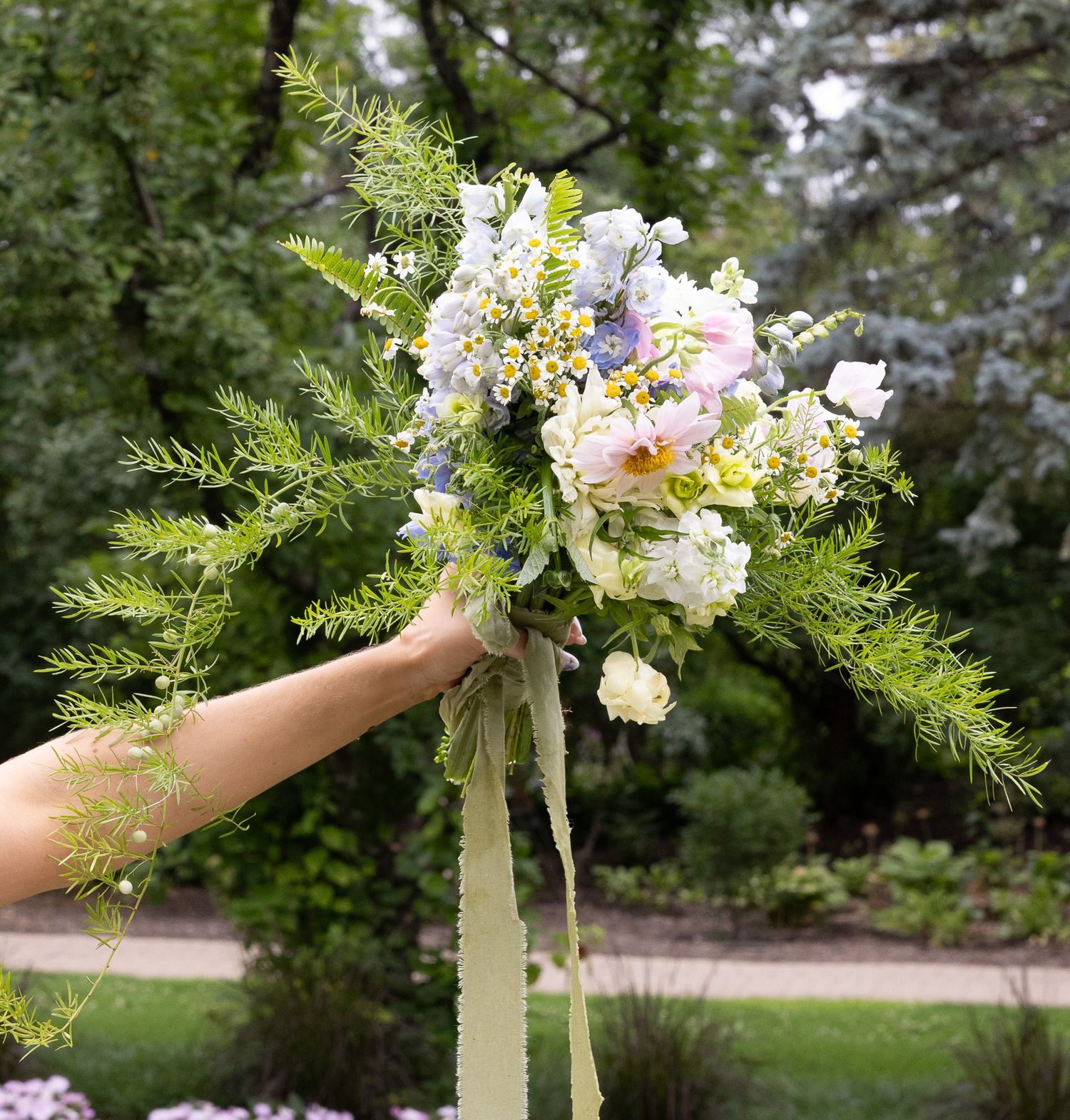 A delicate wedding bouquet with white and pink flowers, ferns, and ribbons held up against a garden background.