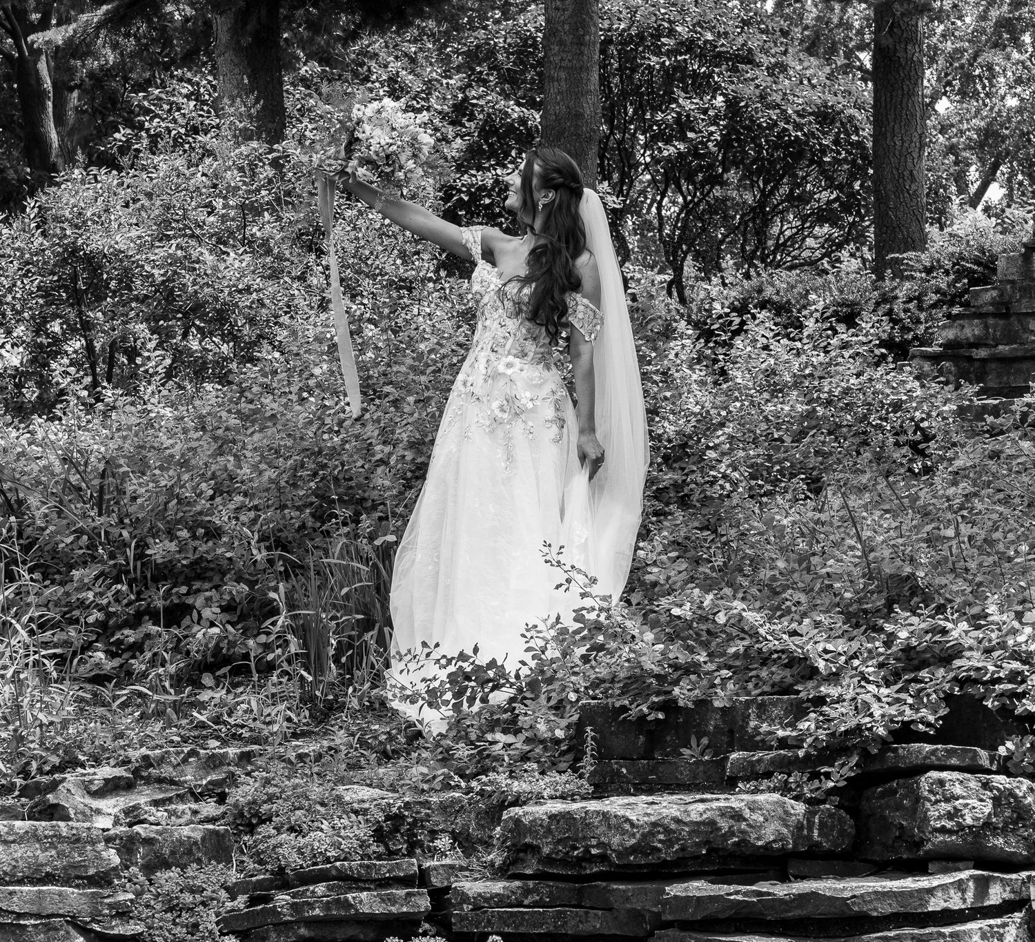 A wedding dress hanging elegantly on a tree branch in a forest setting, captured in black and white photography.
