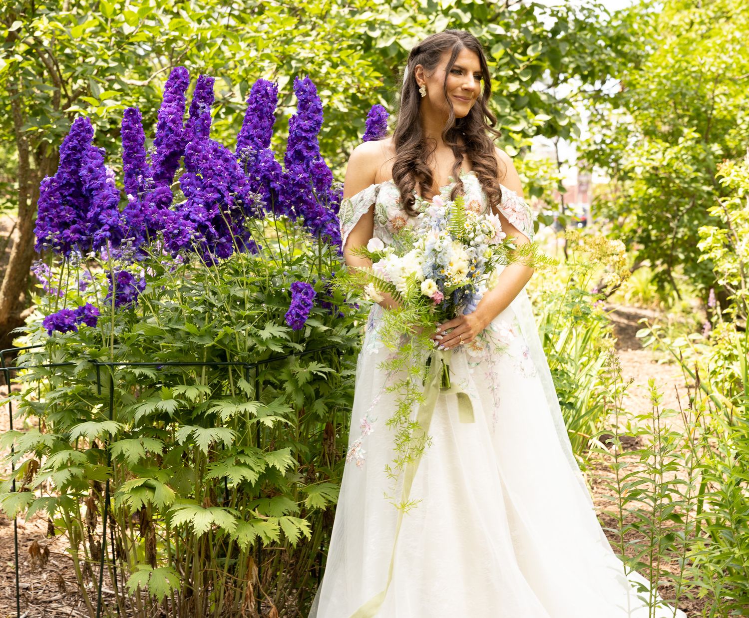 A bride in a white off-shoulder gown holds a floral bouquet while standing next to vibrant purple delphinium flowers in a garden.