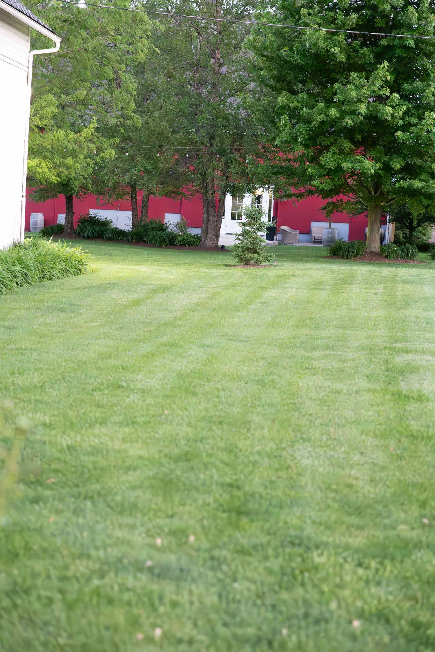 A well-manicured lawn stretches between a house and red outbuildings with mature trees providing shade in the background.