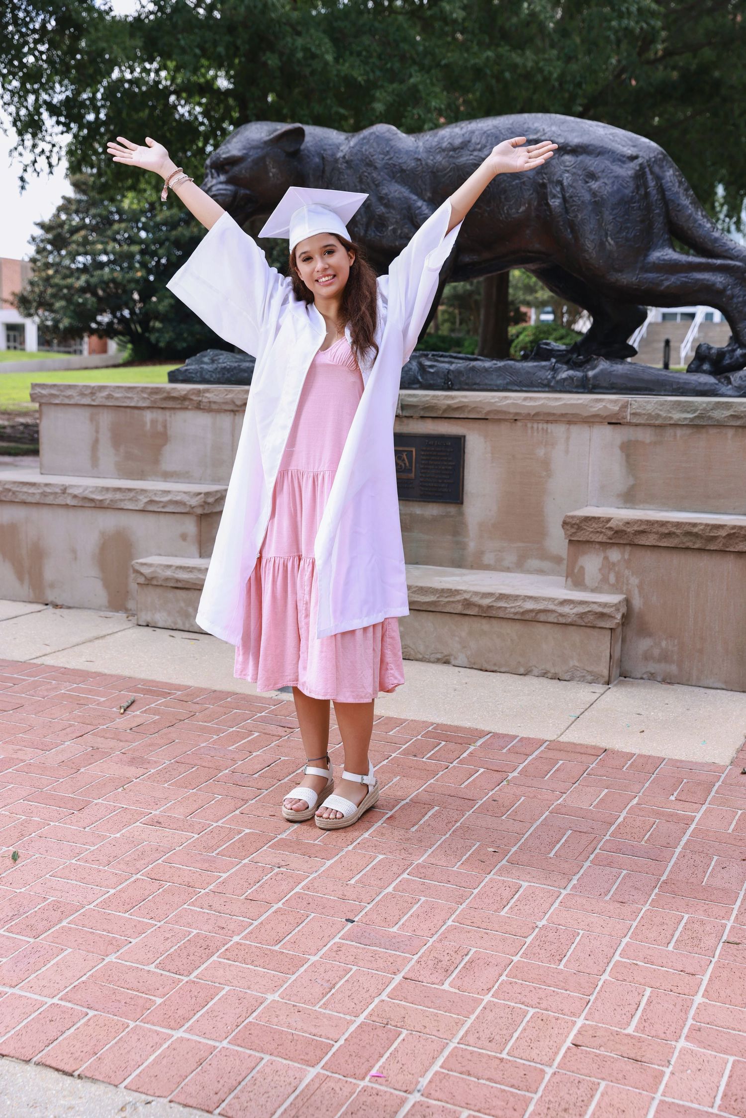 Graduate in white cap and gown celebrating by panther statue on brick plaza wearing pink dress and white slides.