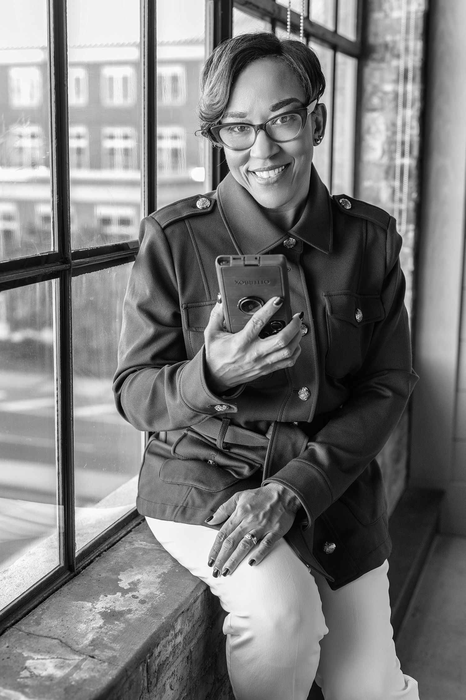 Black and white portrait of someone in glasses and a casual jacket sitting by a window while holding a mobile device.