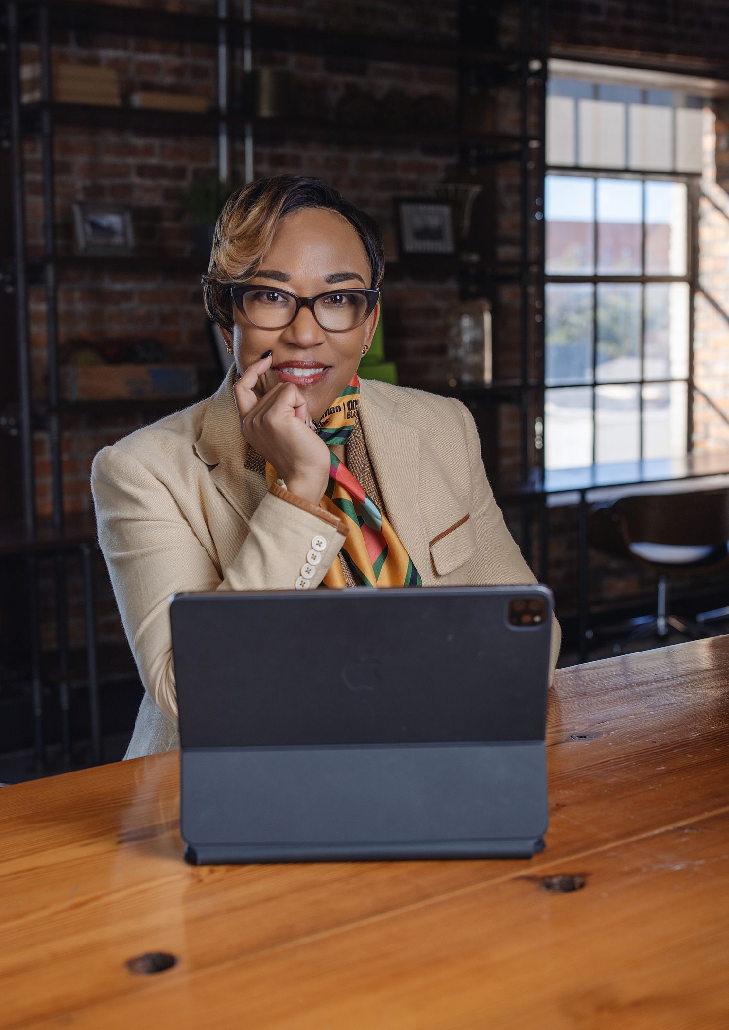Professional wearing a light colored blazer and glasses sits at wooden desk with laptop in modern brick office setting.