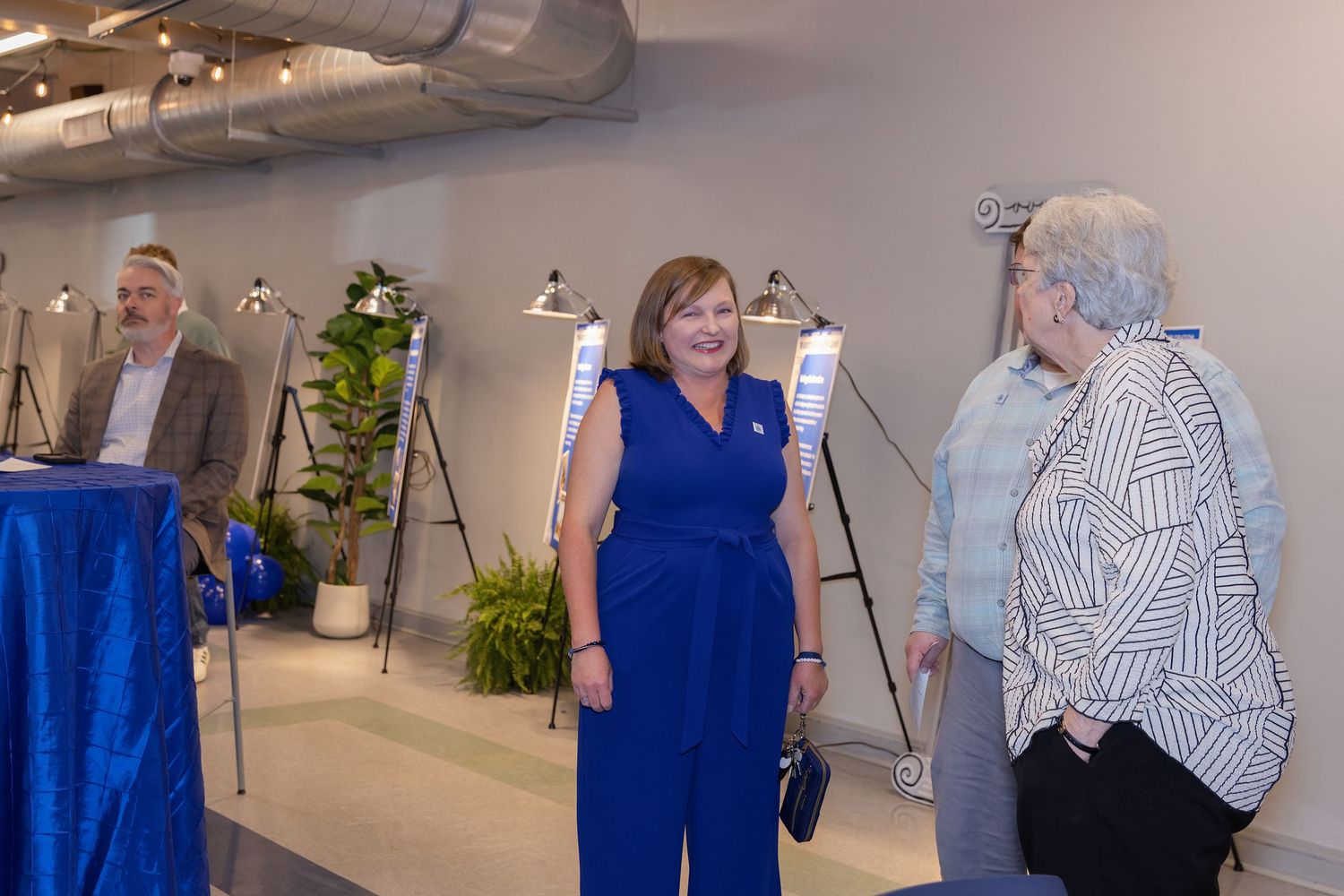 Three people stand in a modern event space with easels, a woman in blue jumpsuit smiling at a guest in patterned blouse.