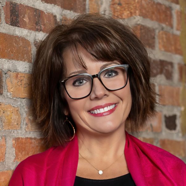 Professional headshot of person in red blazer and glasses against brick wall background.