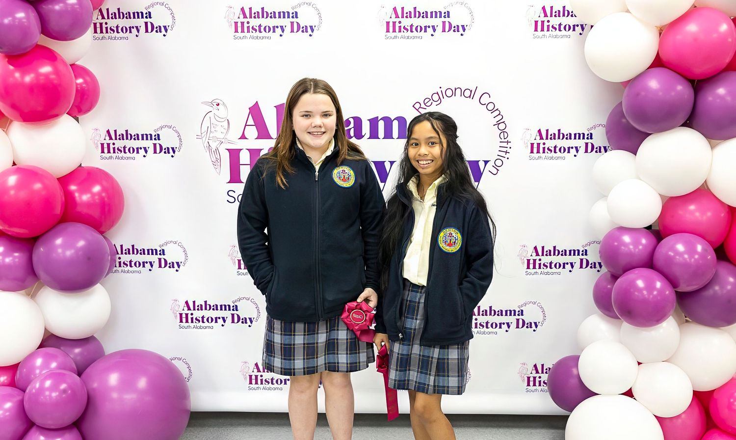 Students posing with awards in front of pink and purple balloon wall at Alabama History Day event.