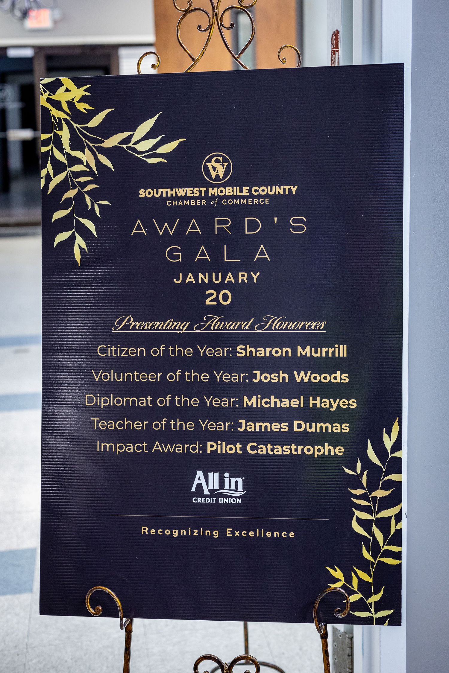 Awards gala signage displays recognition categories and recipients against a dark background with gold leaf accents.