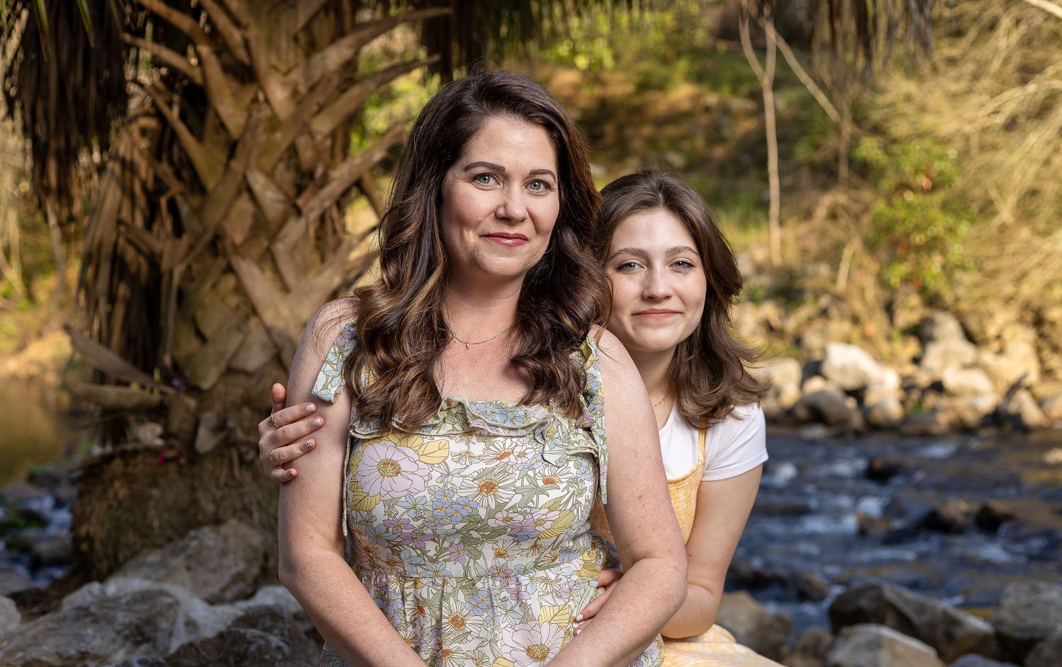 Mother and daughter sit together by a scenic creek with rocks and trees in the background.
