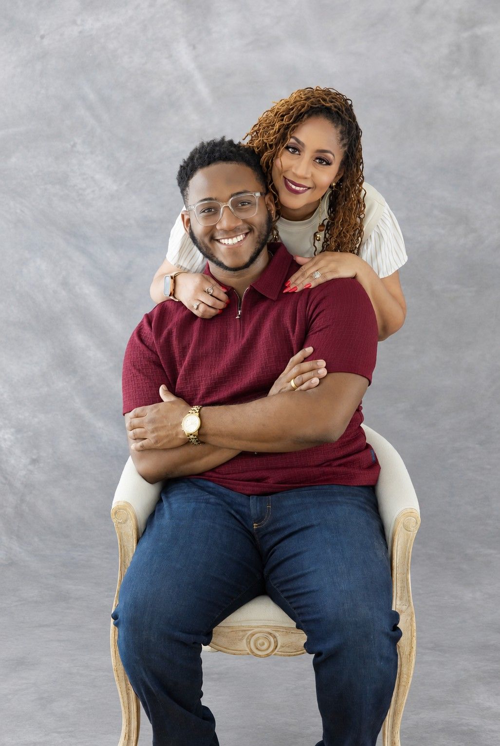 A smiling couple poses warmly in a studio, man seated in chair wearing burgundy polo, woman standing behind him embracing his shoulders.