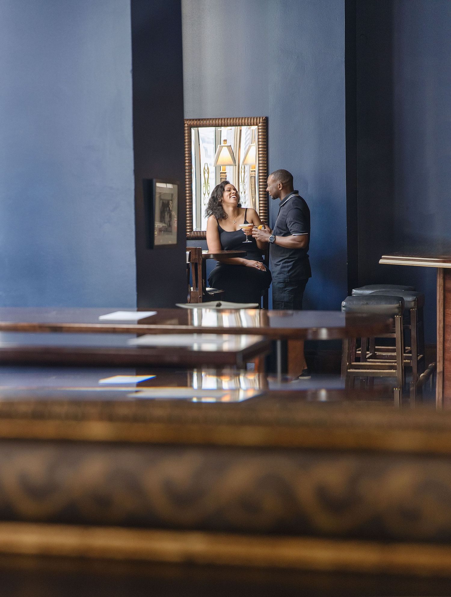 A couple laughing and sharing drinks at an elegant blue-walled bar with ornate wooden furniture.