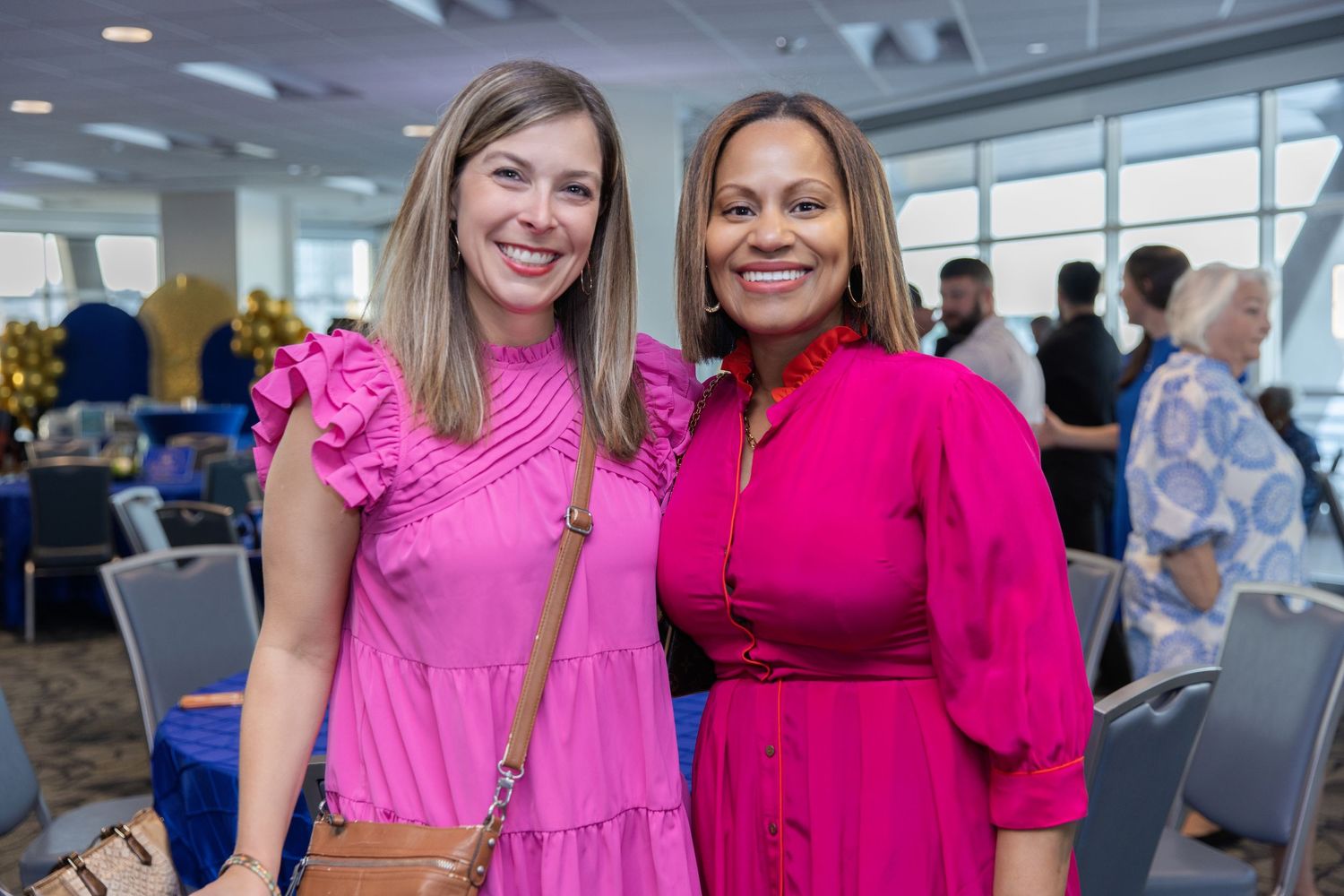 Two women in bright pink dresses smile together at an indoor business or networking event.