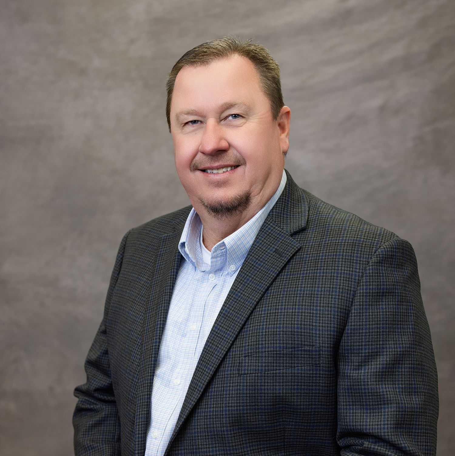 Professional headshot of someone in a dark checkered blazer and light blue shirt against a gray studio backdrop.