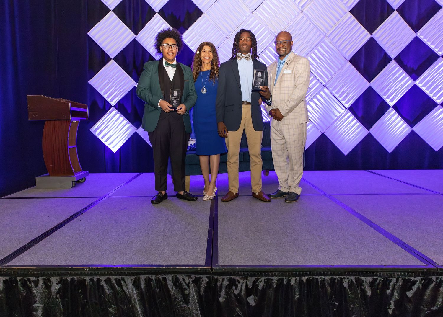Four award recipients pose on stage at a formal ceremony, holding trophies in front of a decorative backdrop.