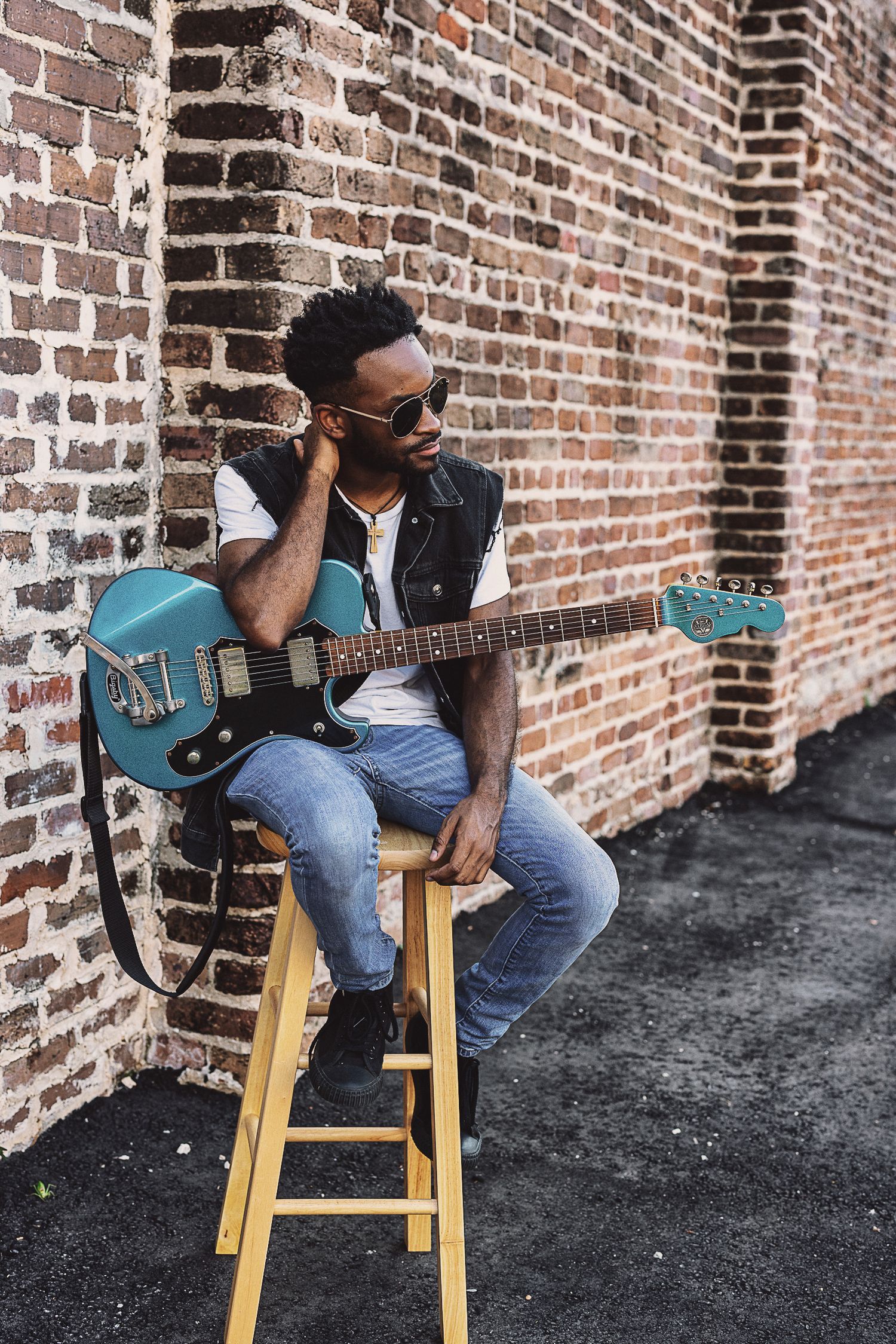 A musician sits on a wooden stool playing a blue electric guitar against a brick wall.