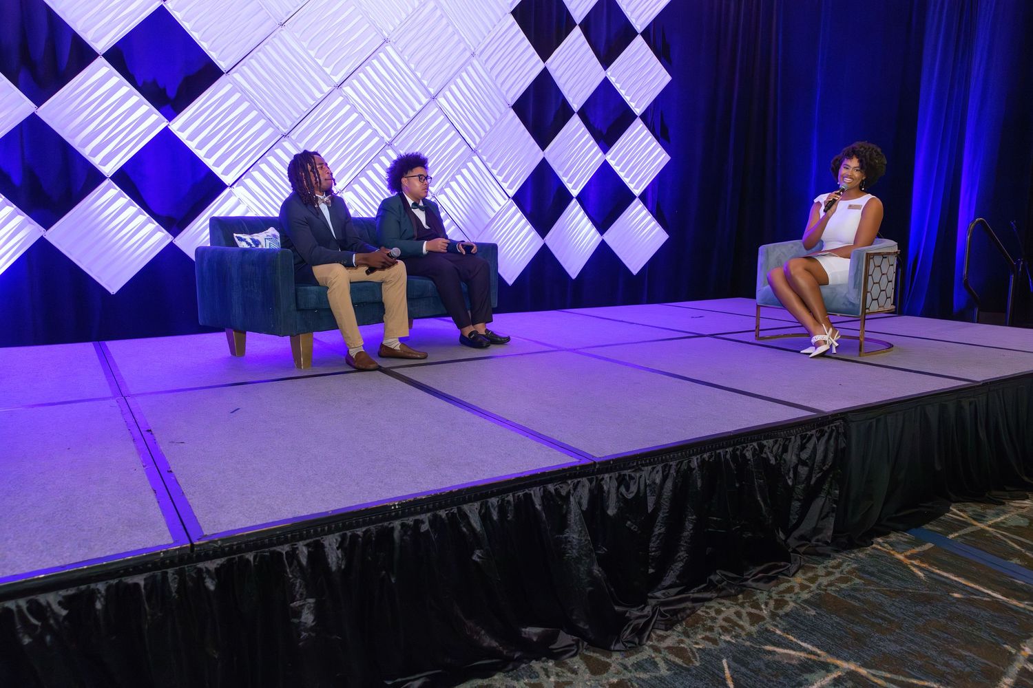 Three people seated on stage at a professional conference with blue and white geometric backdrop lighting.