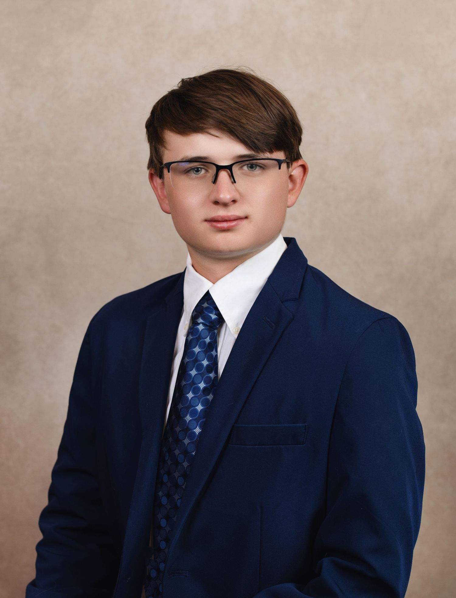 Professional portrait of a student in a navy suit and patterned tie wearing glasses against a neutral backdrop.