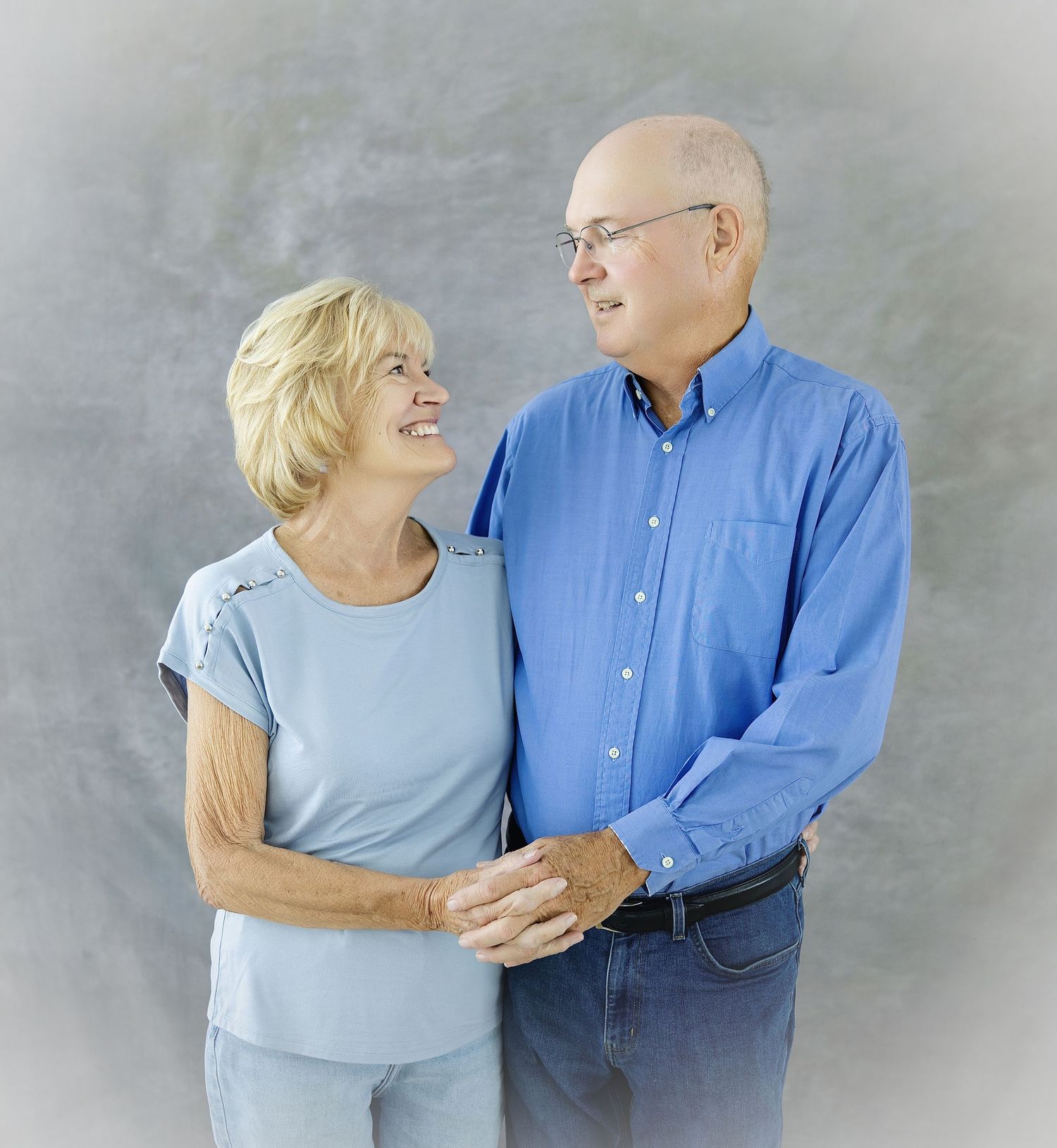 Smiling elderly couple embracing in a studio portrait, woman in light blue top and man in blue shirt with glasses.