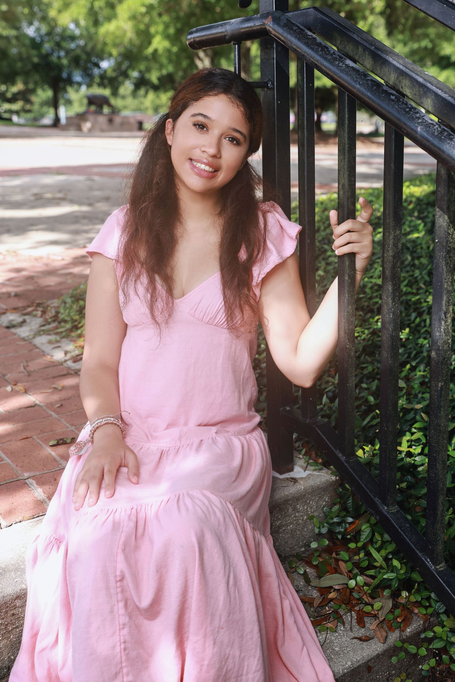 A person in a flowing pink dress sits on outdoor steps next to a black metal railing in a garden setting.