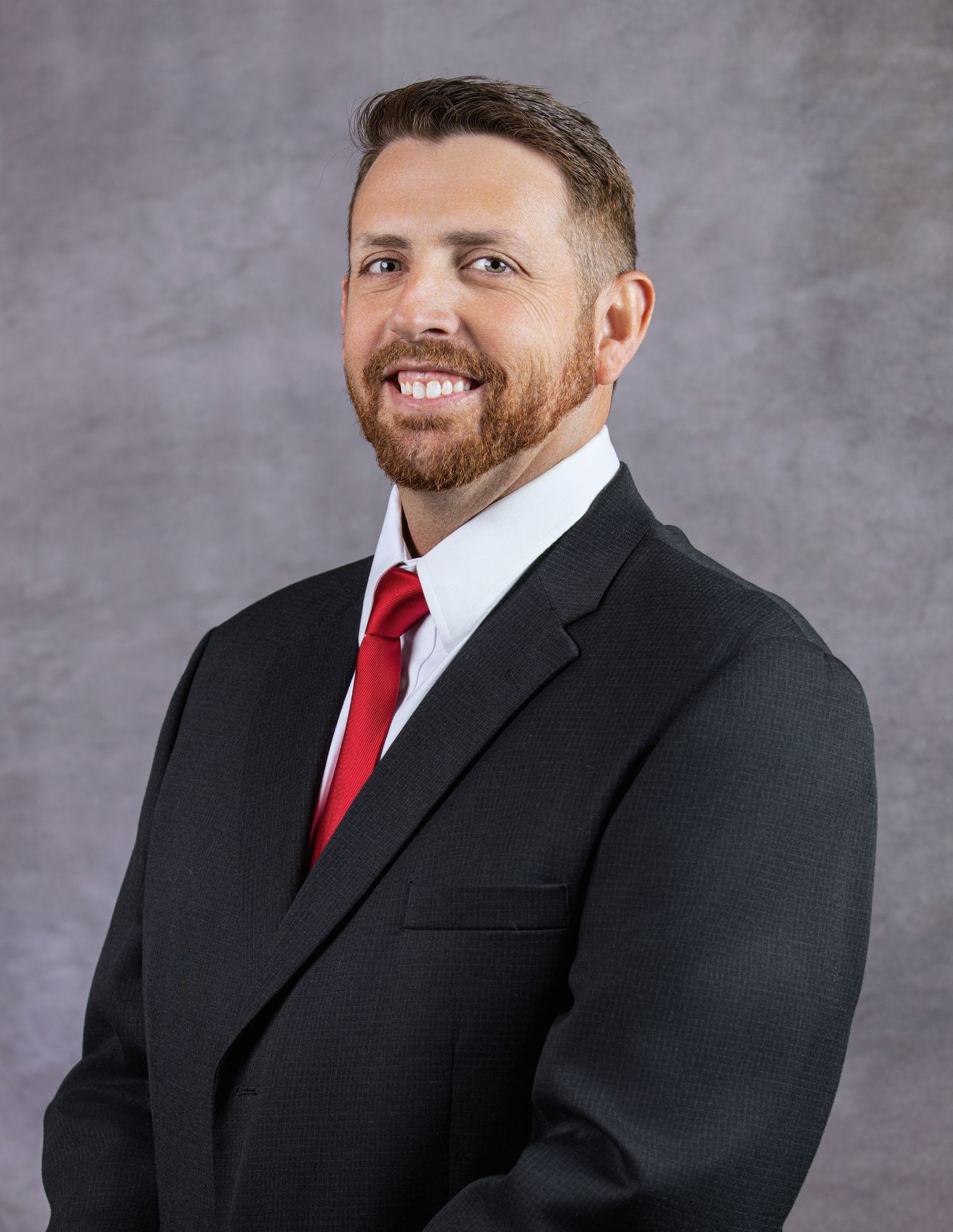 Professional headshot of a smiling man in a black suit and red tie against a gray background.