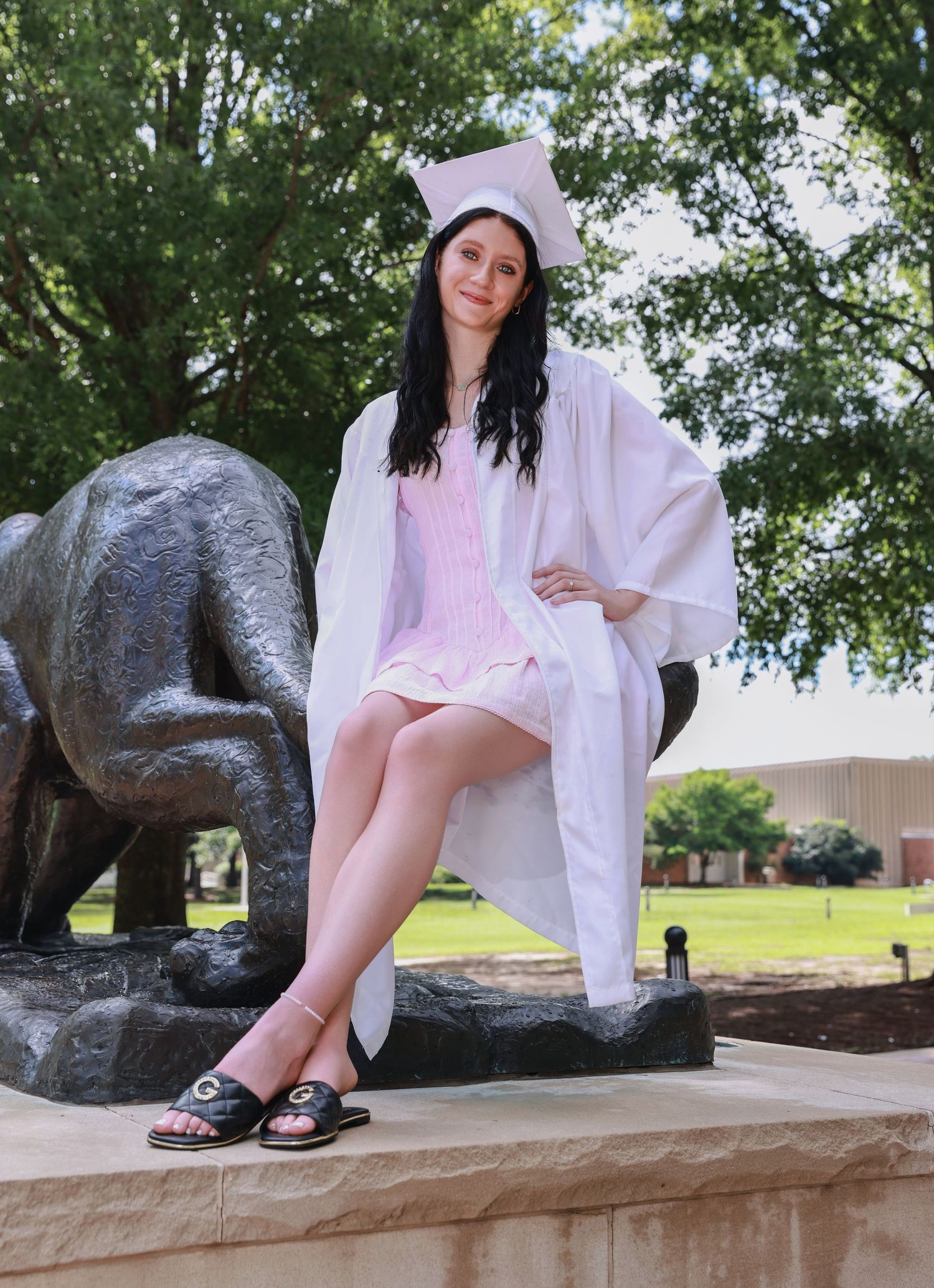 Graduate in white academic regalia sits on stone ledge next to bronze statue in outdoor campus setting.