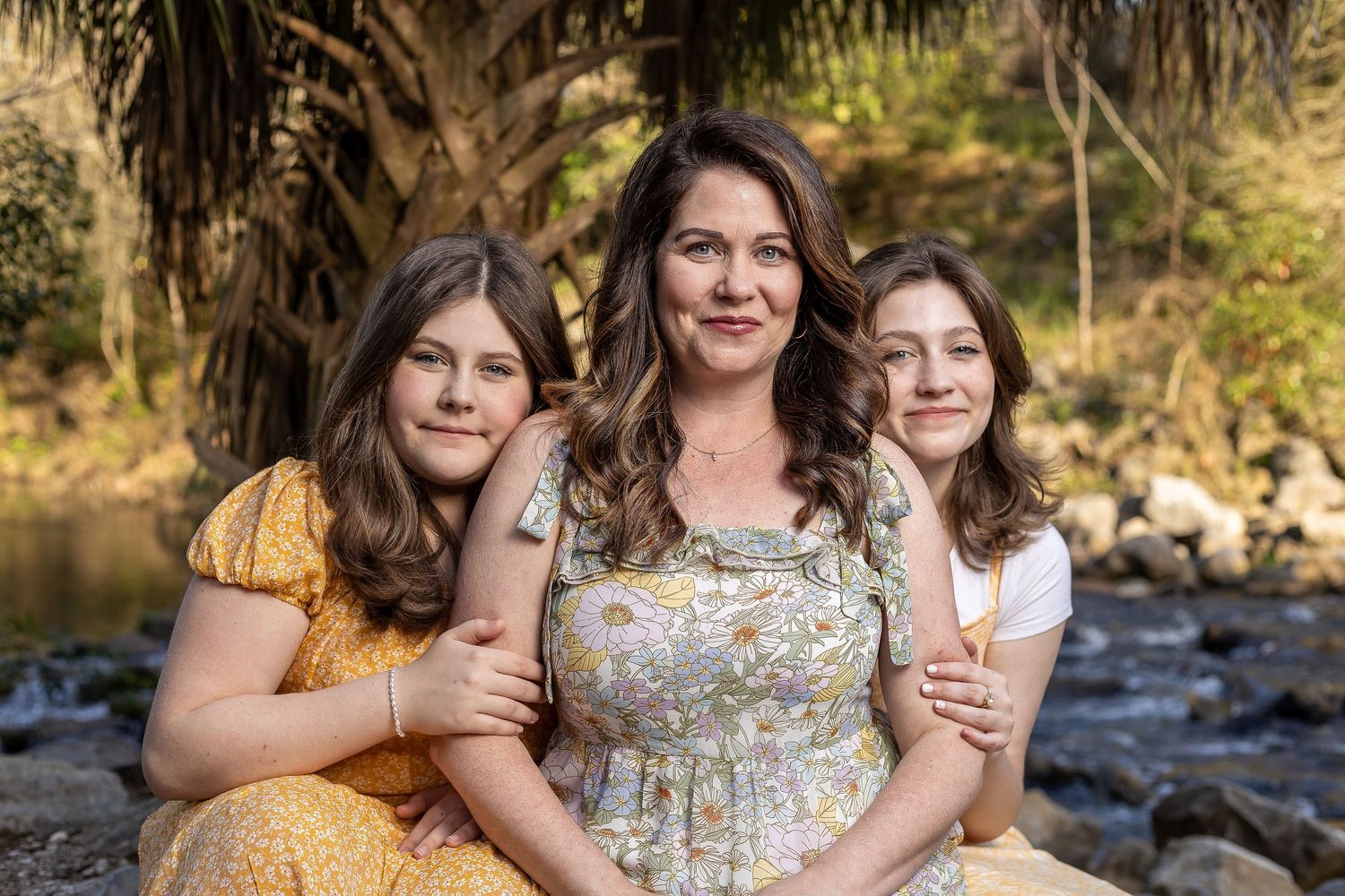 Three women in summer dresses pose together by a creek with rocks and foliage.