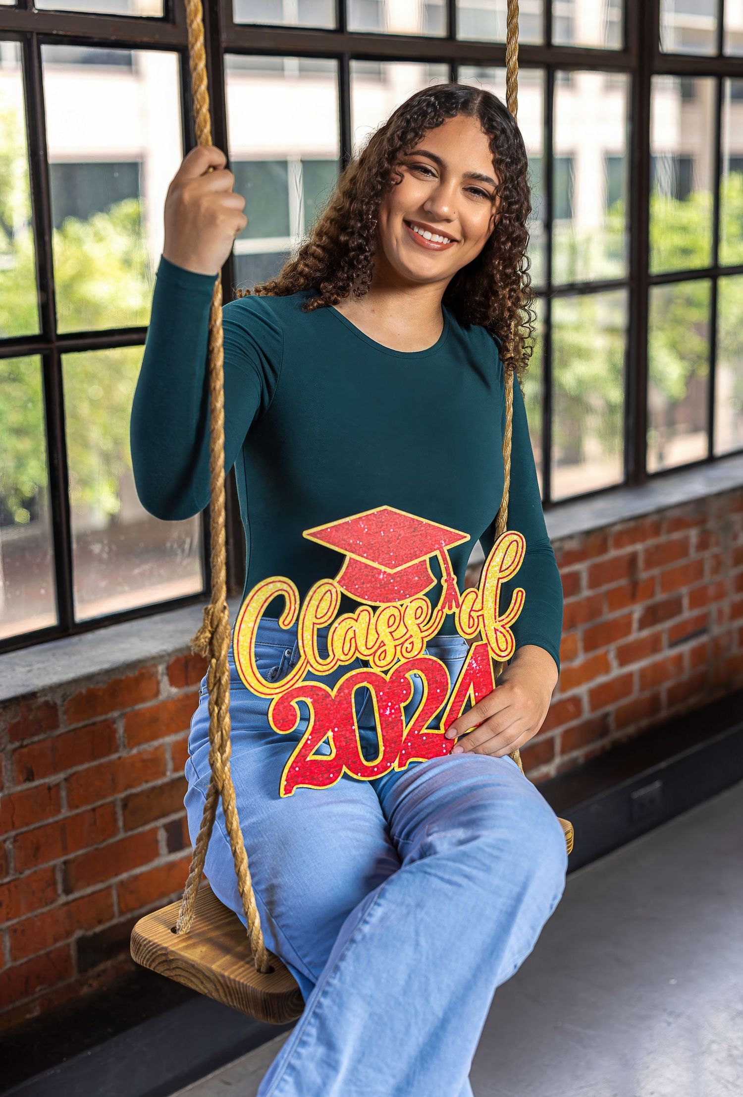 Graduate sits on wooden swing wearing teal sweater with Class of 2024 design and blue jeans.