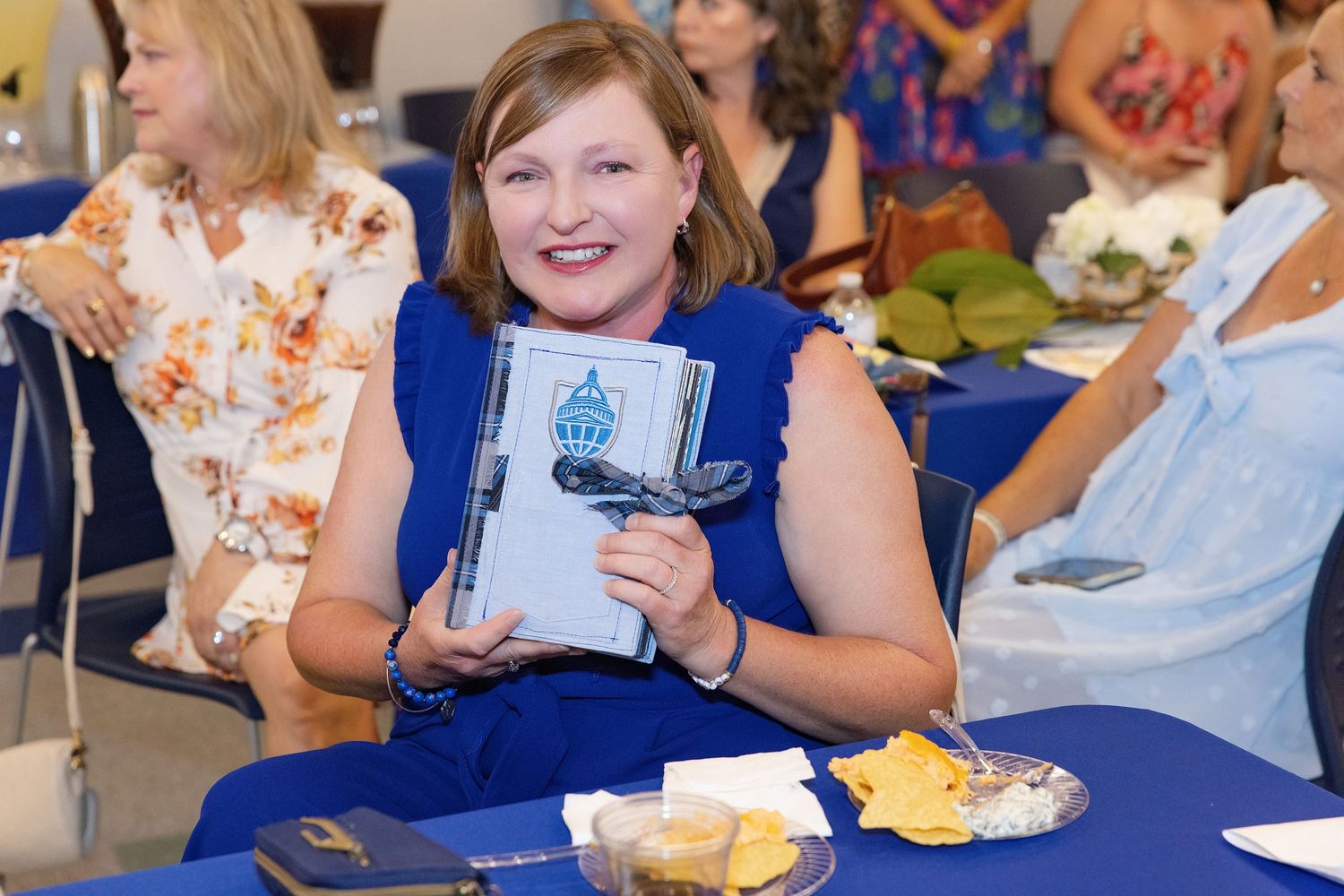 Smiling woman holding a blue book at a banquet table with snacks, surrounded by guests at an indoor event.