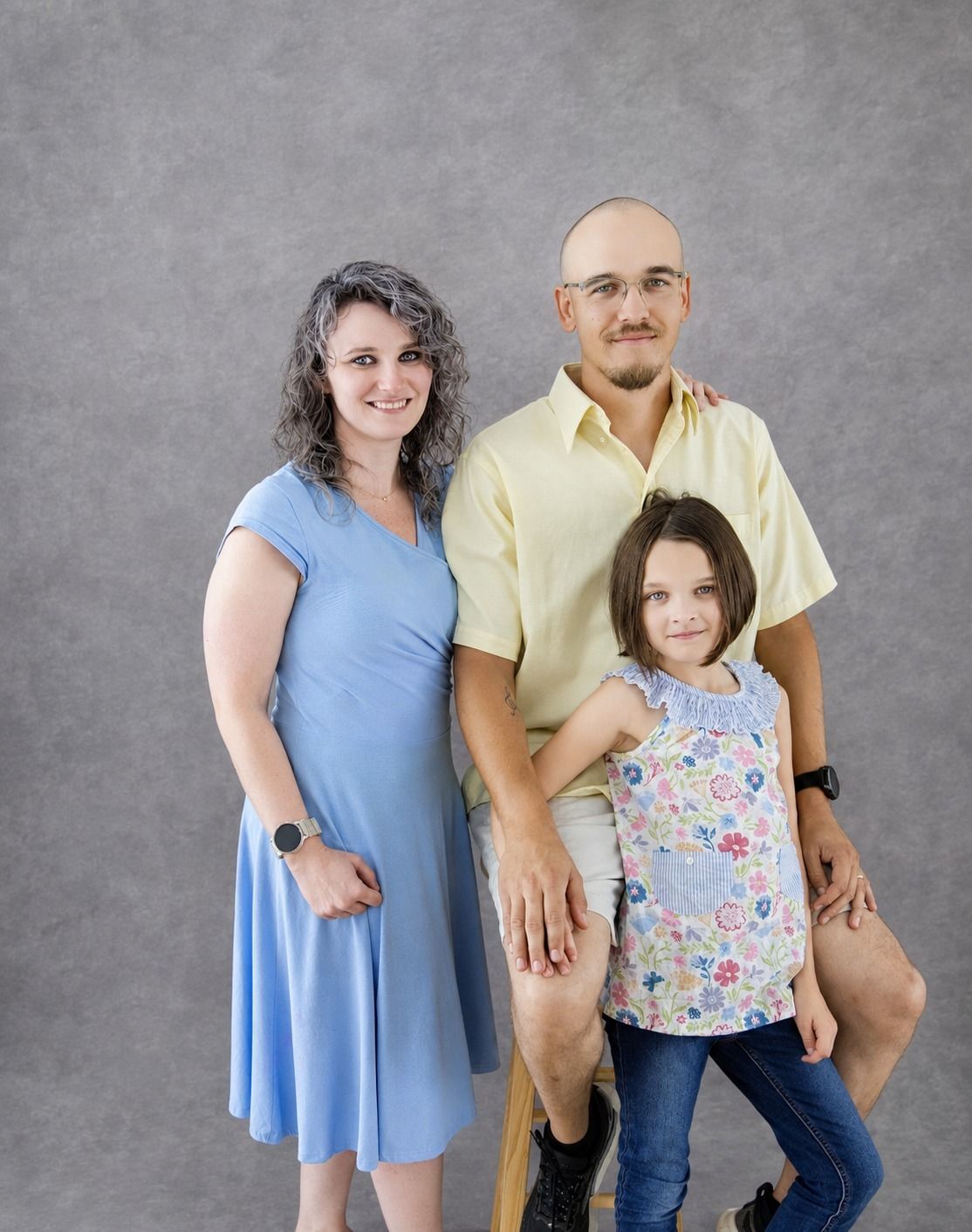 A family of three poses for a studio portrait, featuring a woman in blue dress, bald man in yellow shirt, and young girl.