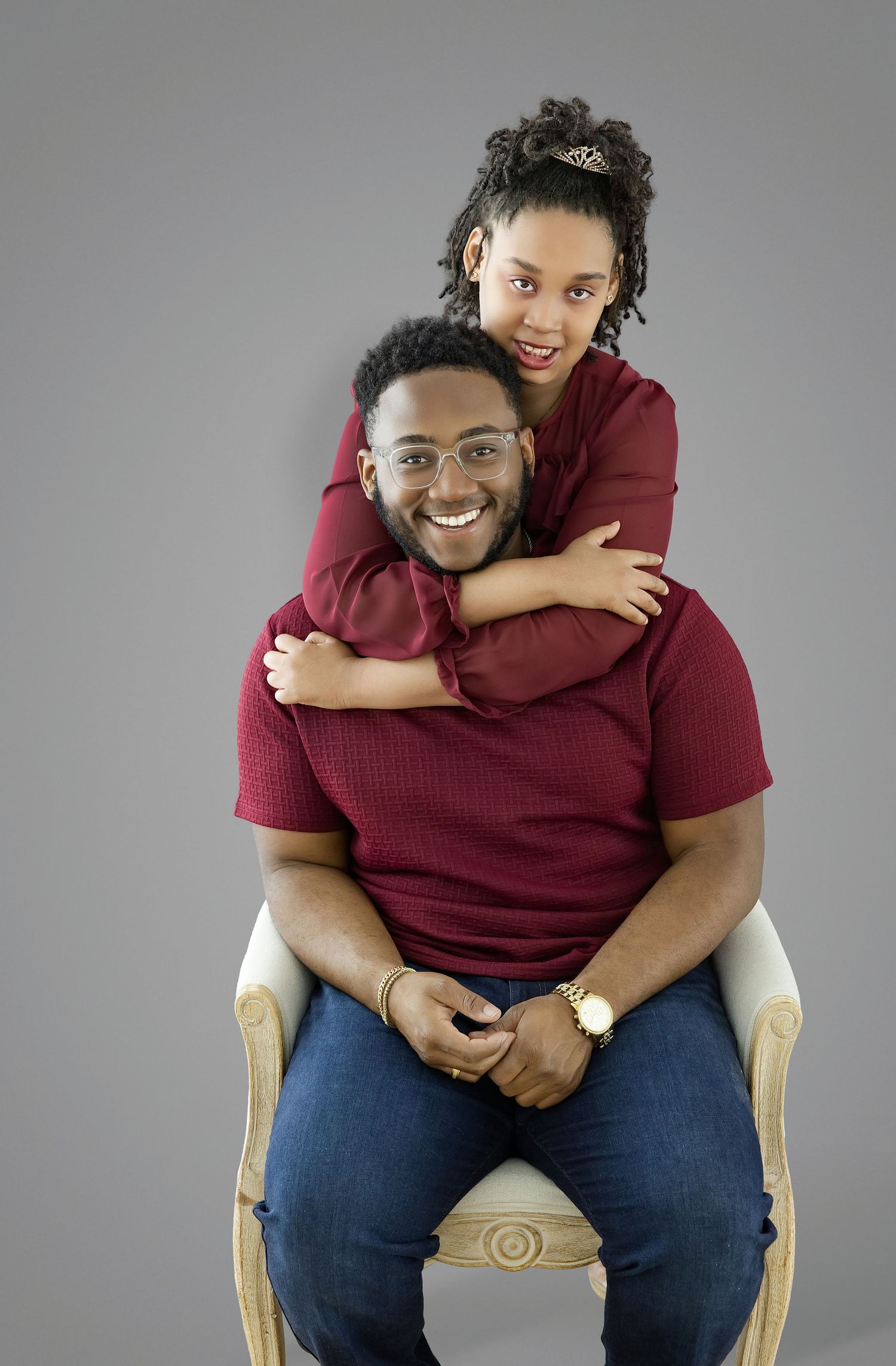 A smiling man in a red shirt sits in a chair while a young woman hugs him from behind, both wearing matching outfits.