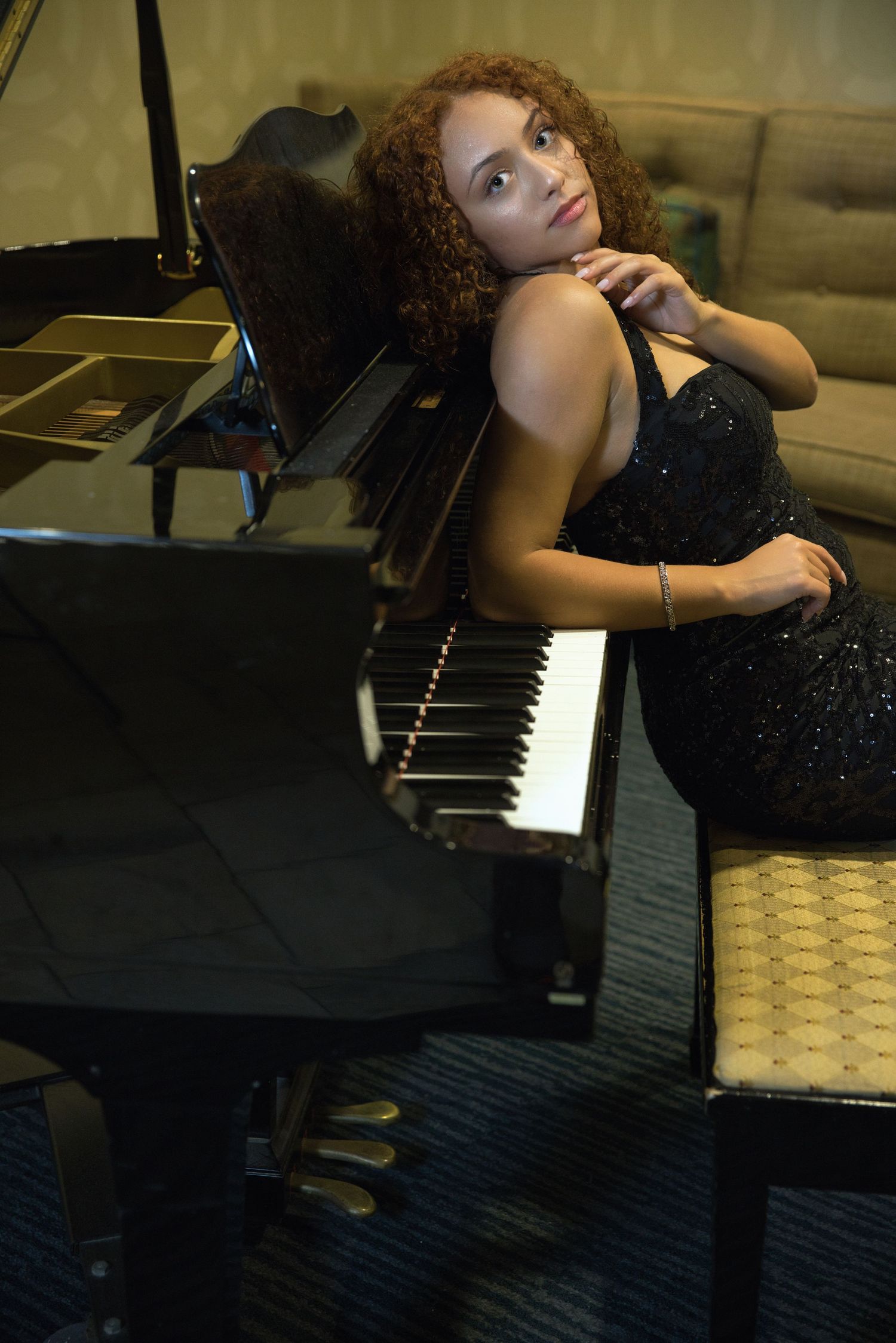 A glamorous woman in a sparkly black dress poses beside a grand piano.