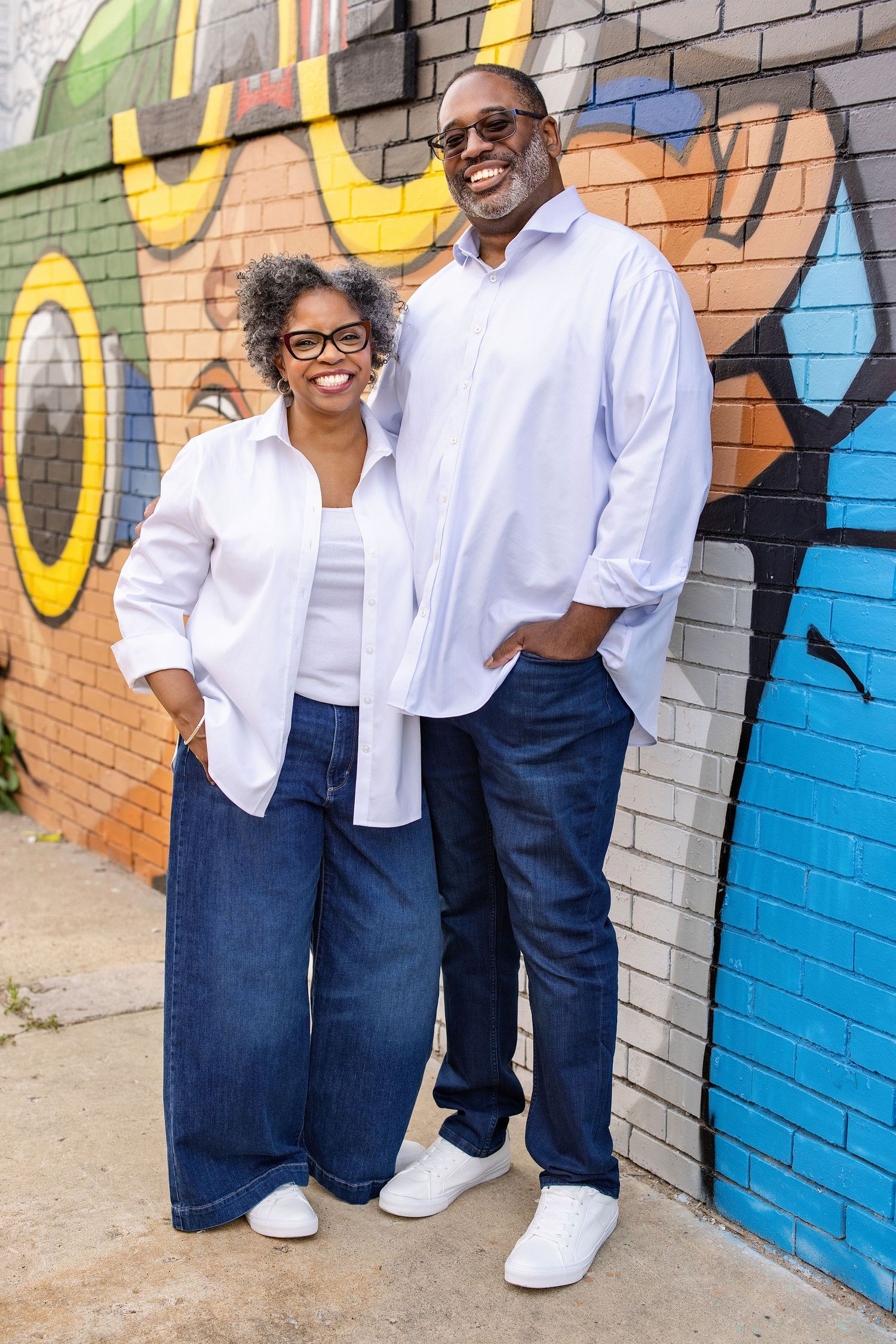 Smiling couple in matching white shirts and jeans posing in front of a colorful graffiti mural on a brick wall.