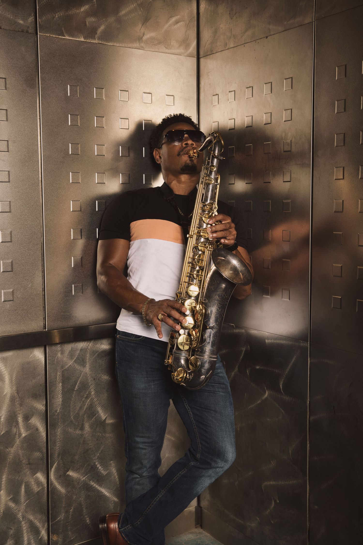 A musician plays saxophone in an elevator with metallic textured walls.
