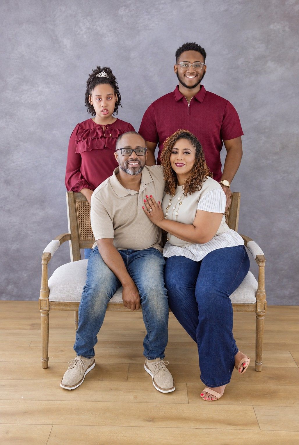 Family portrait of four in burgundy and neutral outfits, with two parents seated on a cushioned bench and two children standing behind them.