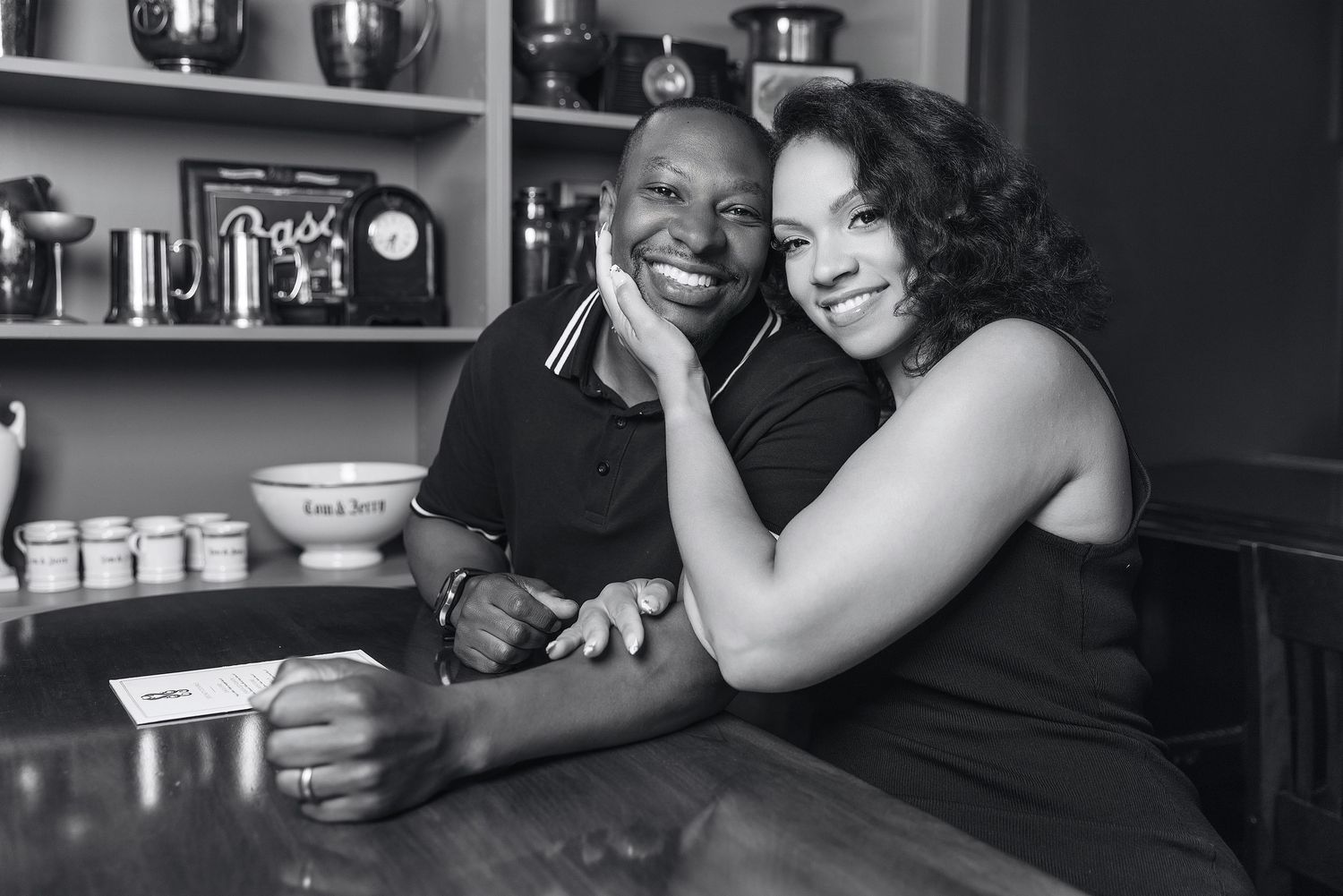 Black and white photo of a smiling couple posing together at a restaurant counter, woman leaning on mans shoulder.