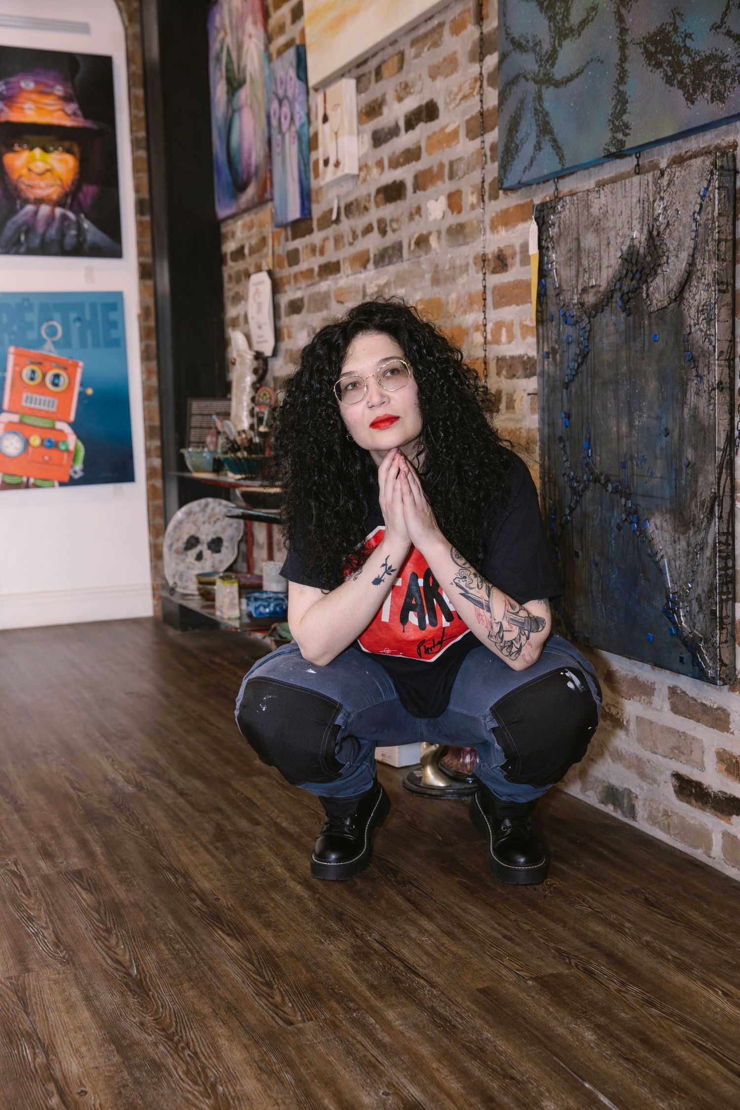 Alternative style portrait with subject in crouching pose against exposed brick wall with artwork displayed.